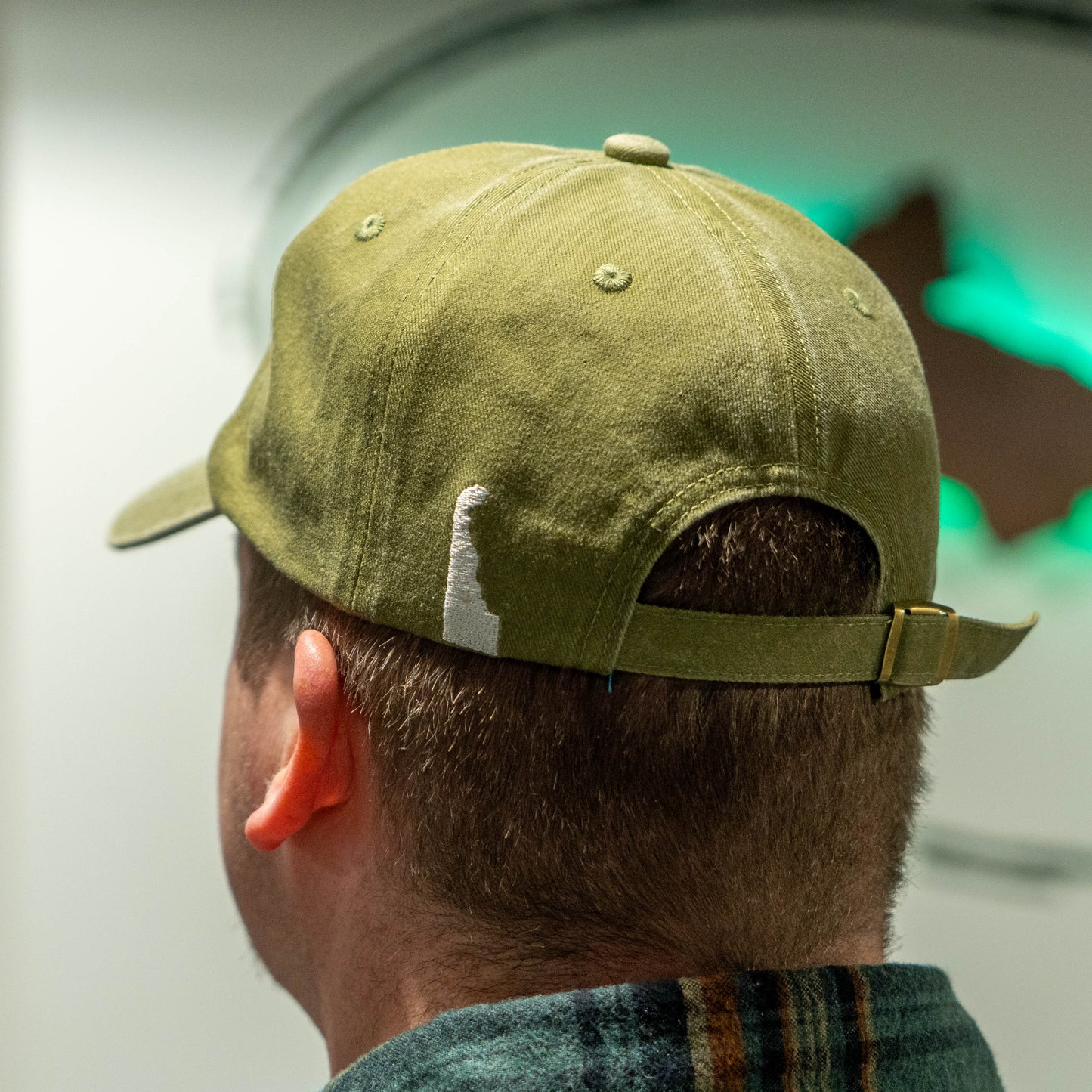 Backside view of man wearing an olive colored dad hat with a white embroidered shape that represents the state of Delaware on the back left side of the hat