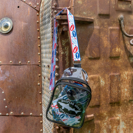 A clear Merchtank bag from the Drink Summer Louder Festival Kit, featuring a vibrant red, blue, and white strap, hangs from a rusted metal surface with bolts and a round window—ideal for carrying your festival essentials this summer.