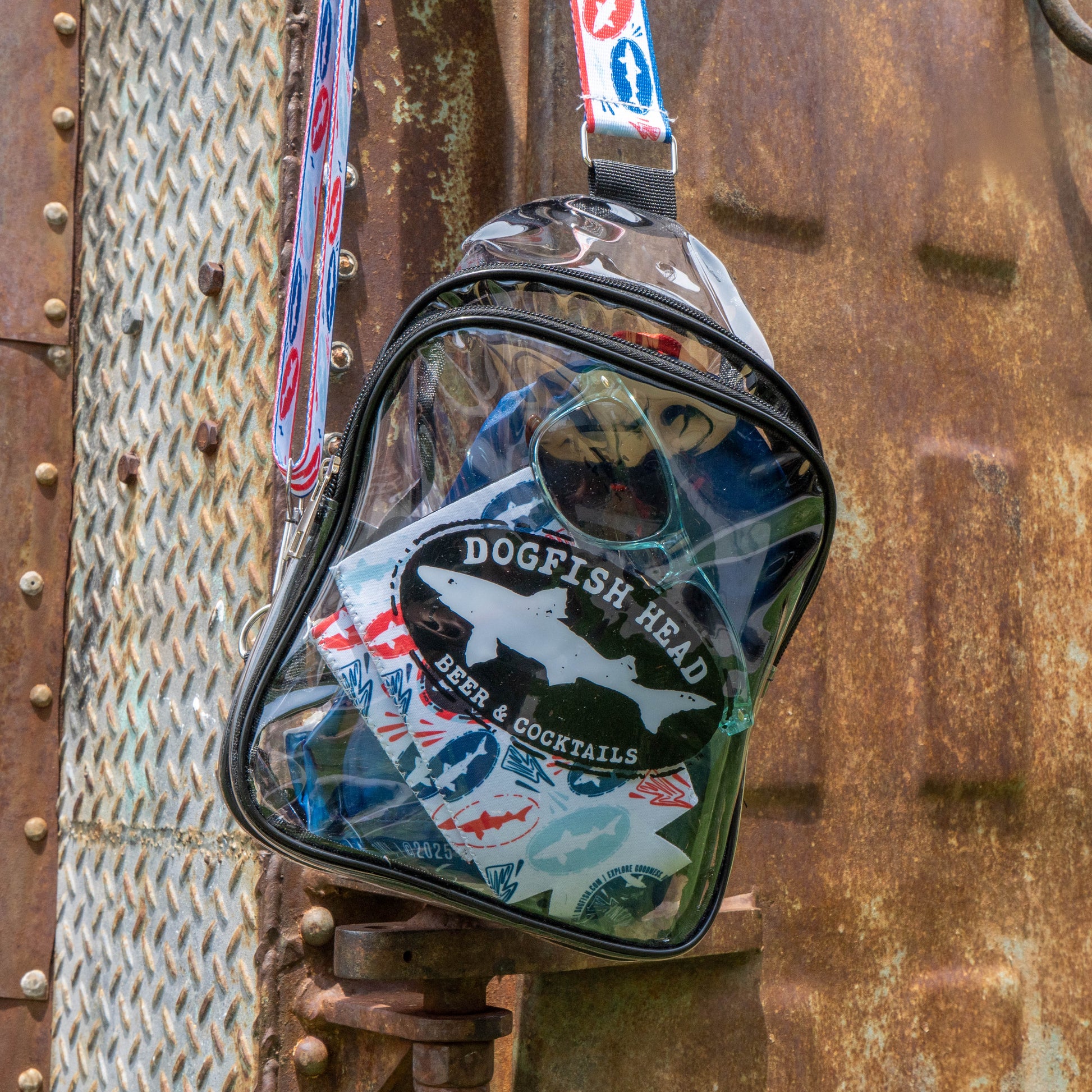 A clear plastic backpack with a Merchtank Drink Summer Louder Festival Kit patch hangs on a rusty metal surface. With vibrant straps and visible contents, it's your go-to kit for summer festival essentials.