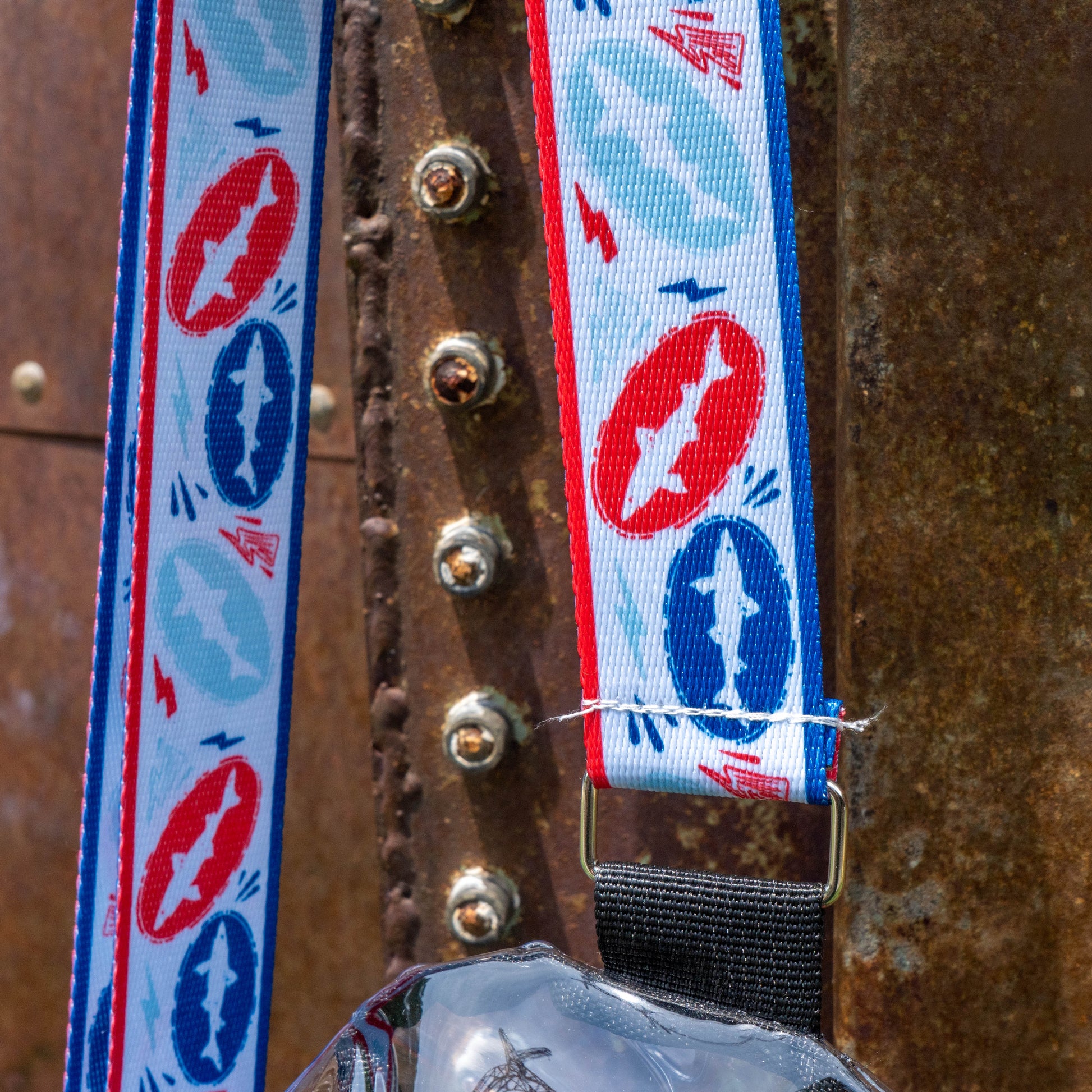 A close-up of the Merchtank Drink Summer Louder Festival Kit lanyard, featuring red, blue, and white airplane and star designs—an ideal summer festival accessory—hanging from a hook on rustic metal.