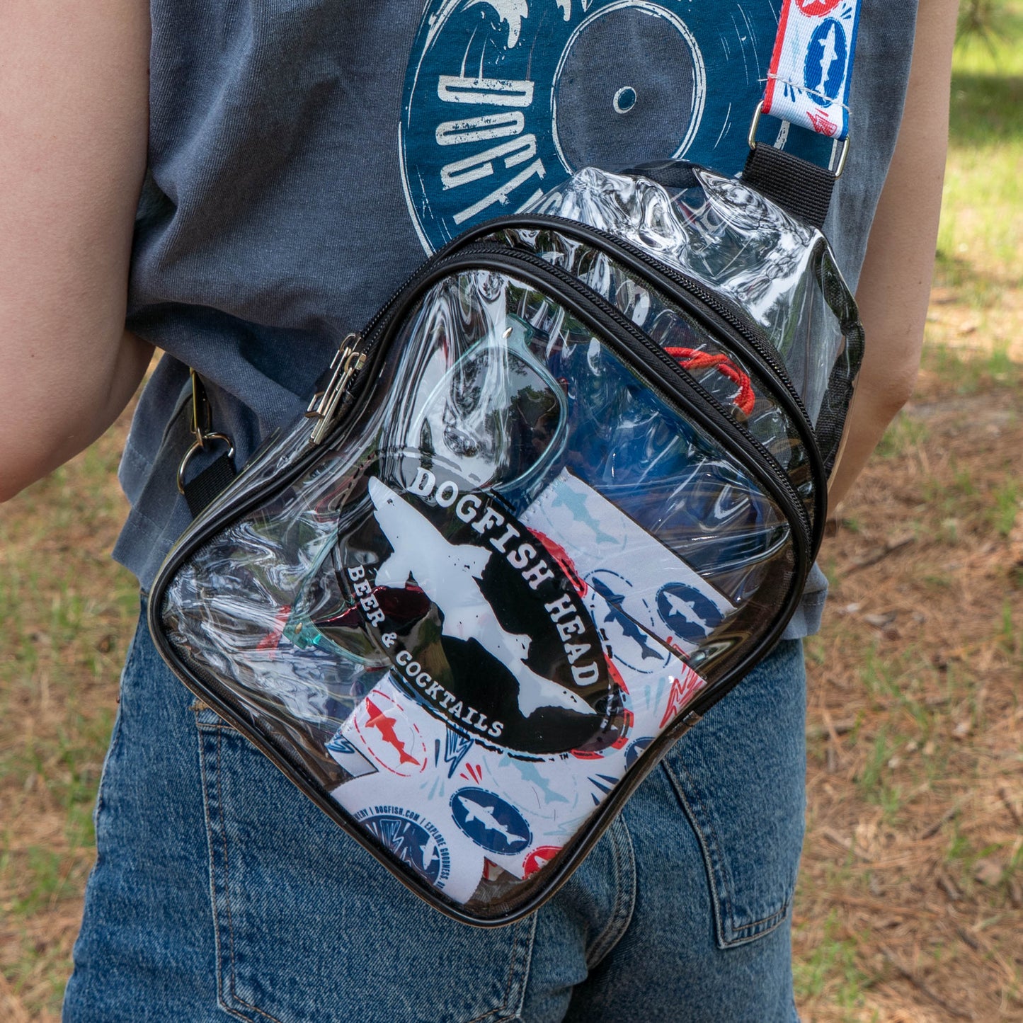 Wearing blue jeans and a blue graphic tee, someone carries a clear crossbody bag with a Merchtank Drink Summer Louder Festival Kit—complete with stickers and a can—for perfect summer festival vibes on the grass.