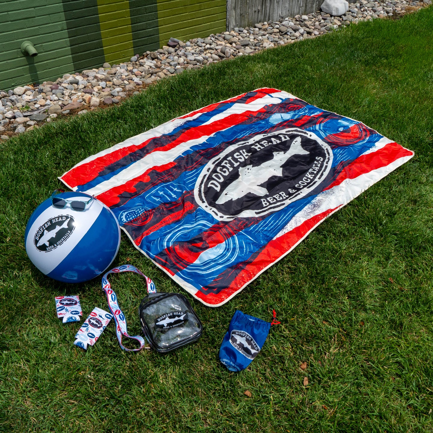 The Merchtank Drink Summer Louder Festival Kit, featuring essentials like a beach ball, blanket, drawstring bag, sunglasses, and lanyards, is displayed on grass—perfect for celebrating summer festivals in style.