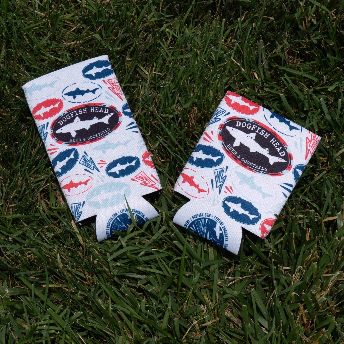 Two white drink koozies with red, blue, and teal shark designs and "Dogfish Head Beer & Cocktails" logos rest on grass—summer must-haves from Merchtank’s Drink Summer Louder Festival Kit.
