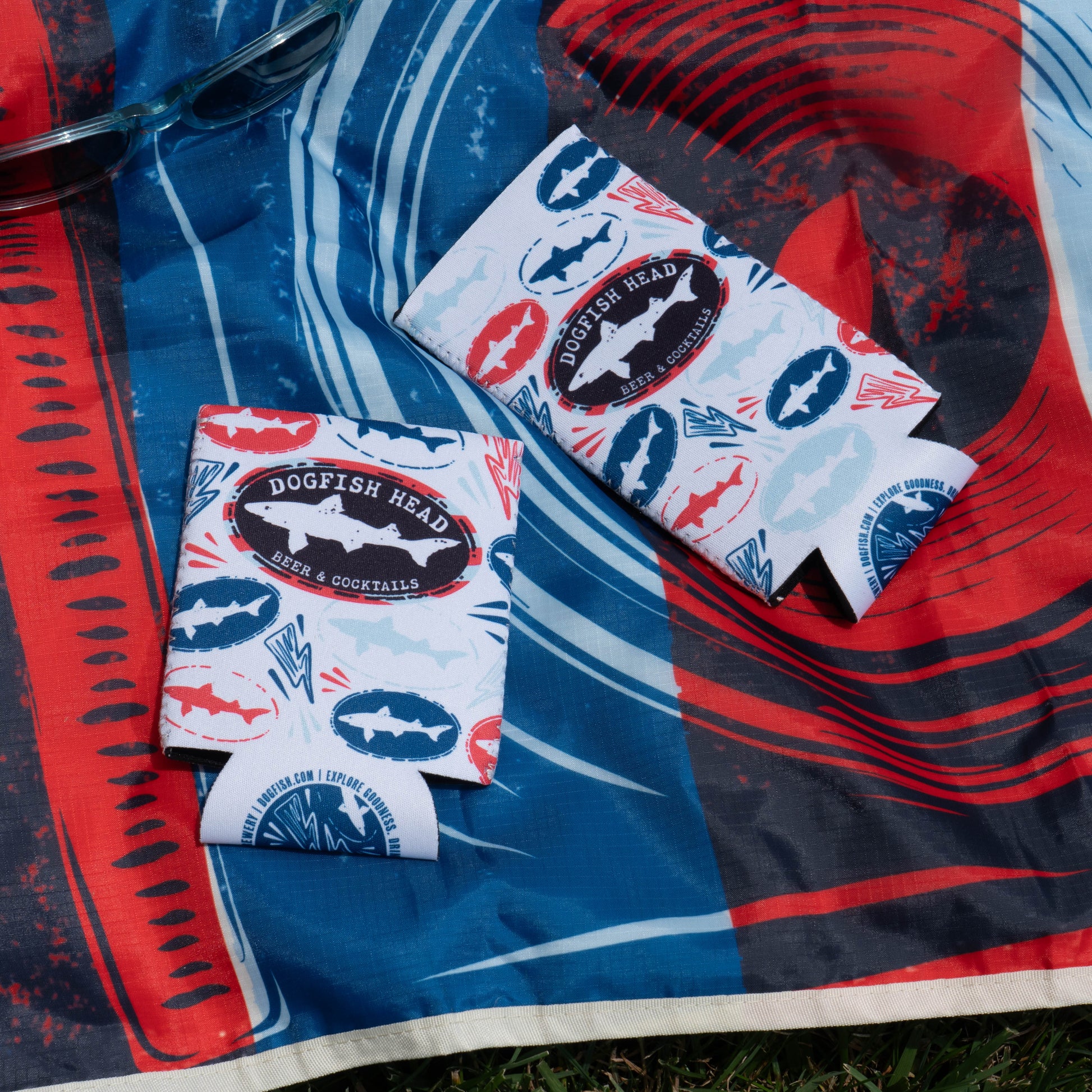 Two Merchtank Drink Summer Louder Festival Kit can coolers with blue and red fish patterns sit on a red, blue, and white cloth outdoors—perfect festival essentials. Sunglasses are partially visible in the upper left corner.