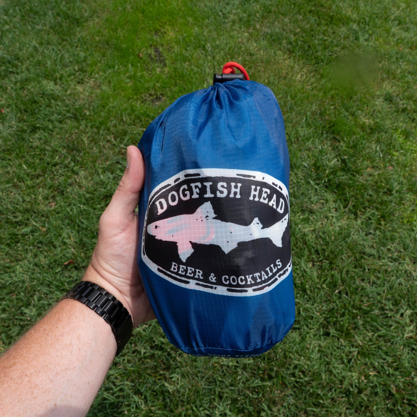 A hand holds a blue Drink Summer Louder Festival Kit drawstring bag by Merchtank, ideal for festival essentials, with a Beer & Cocktails logo, on green grass. The person is wearing a black wristwatch.