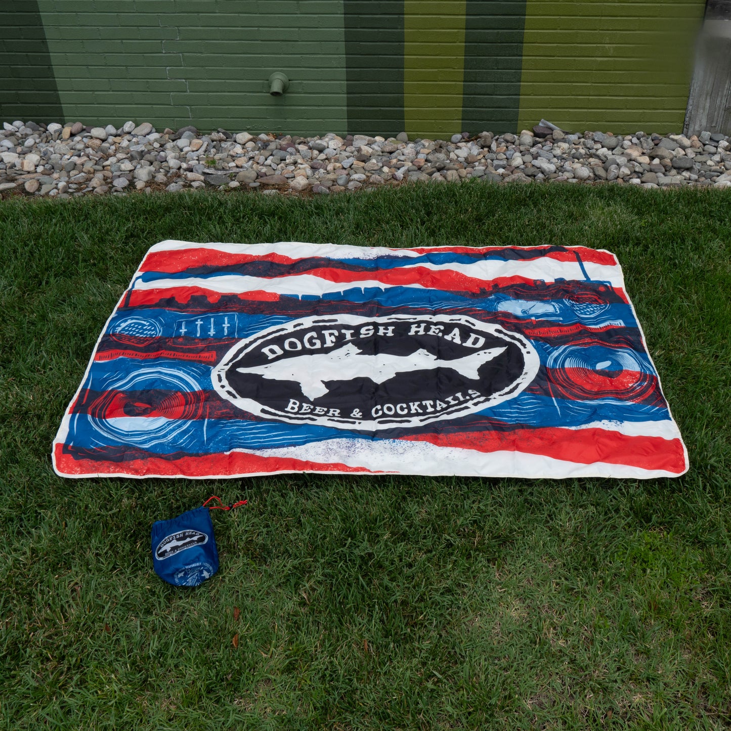 A vibrant blanket from the Merchtank Drink Summer Louder Festival Kit lies on grass by a building, ready for festival moments. It includes a matching blue carrying pouch. Rocks line the background at the base.