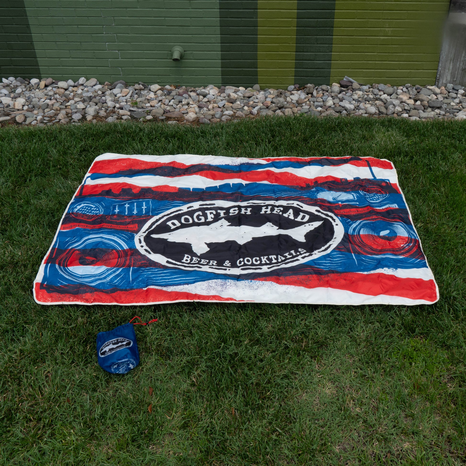 A vibrant blanket from the Merchtank Drink Summer Louder Festival Kit lies on grass by a building, ready for festival moments. It includes a matching blue carrying pouch. Rocks line the background at the base.