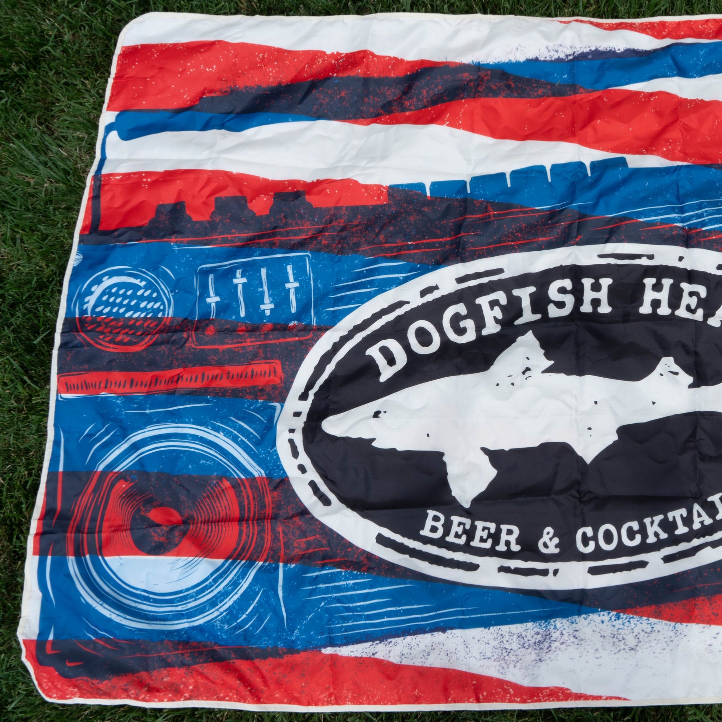 A vibrant outdoor blanket on grass, with red, white, and blue stripes and a bold dogfish shark logo reading “Dogfish Head Beer & Cocktails”—the Merchtank Drink Summer Louder Festival Kit: your must-have for summer fun!.