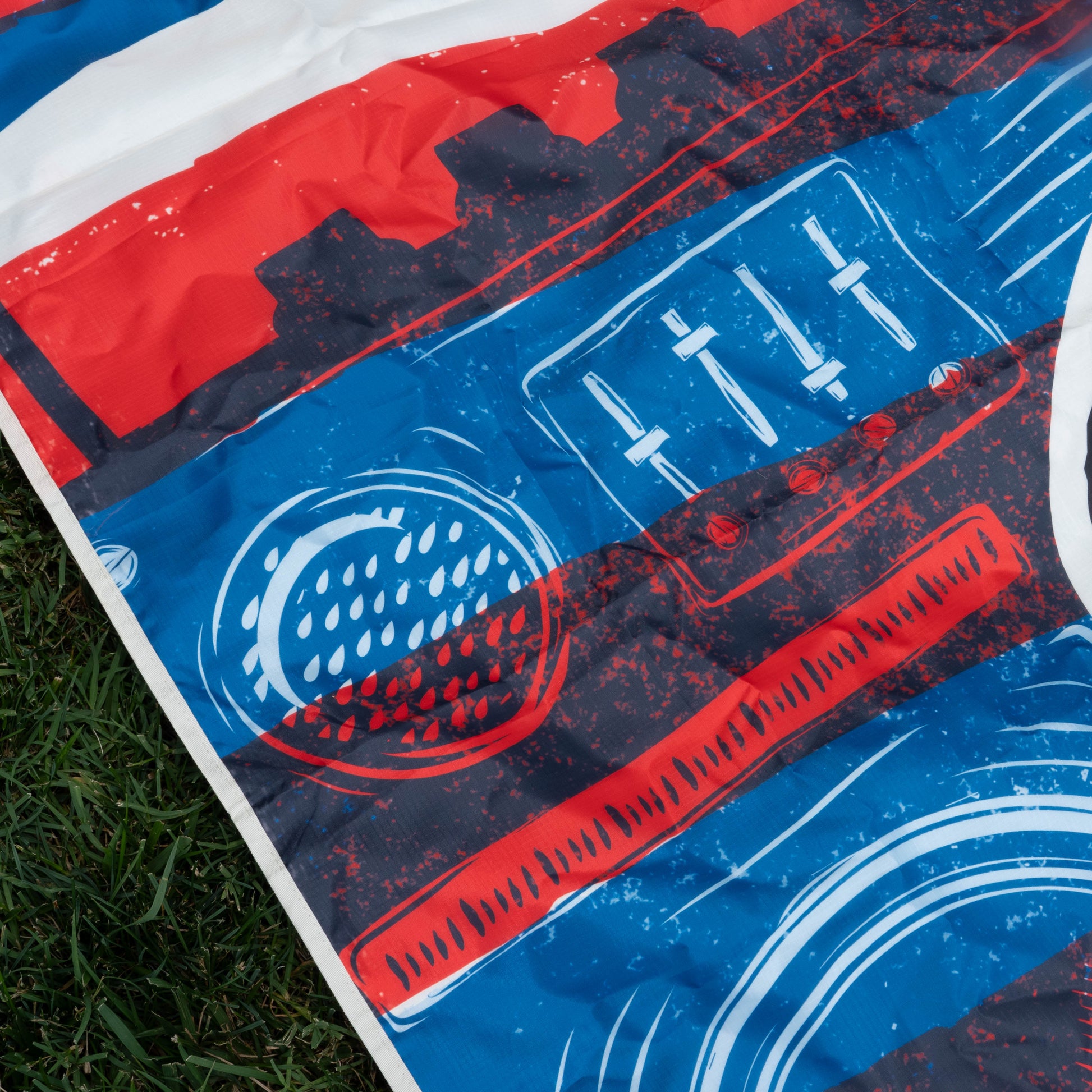 A vibrant fabric featuring a retro boombox in red, blue, and white lies on green grass—a festival essential from the Drink Summer Louder Festival Kit by Merchtank. The design includes detailed dials, sliders, and speakers.