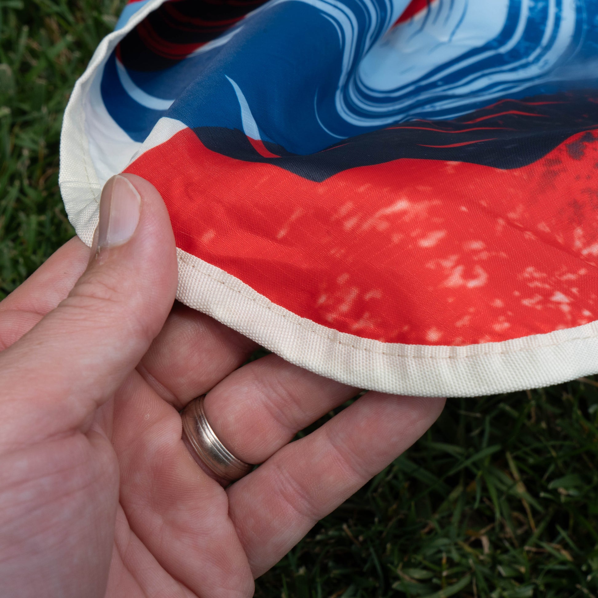 A close-up of a hand with a silver ring holds vibrant red, blue, and white fabric beside green grass—a great way to show off your summer must-haves with the Drink Summer Louder Festival Kit by Merchtank.