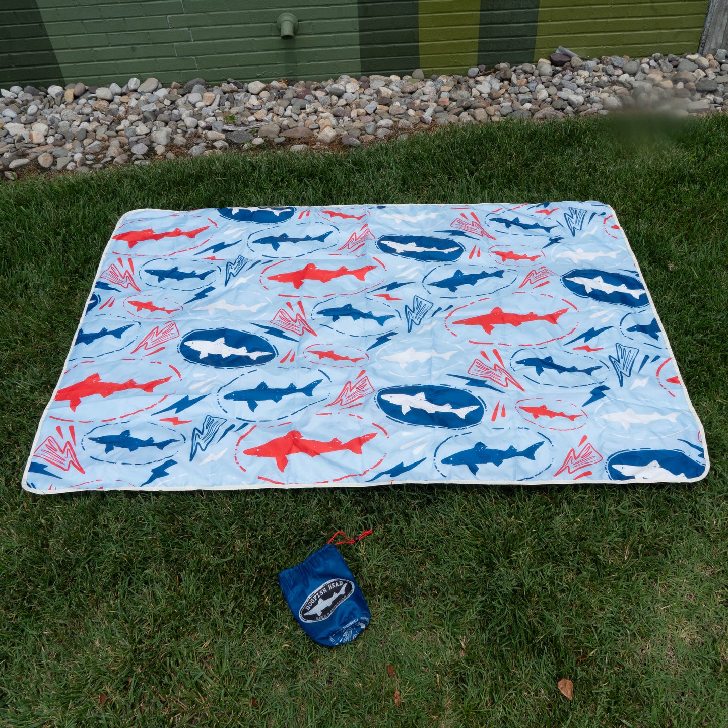 A blue and white picnic blanket with red and blue shark patterns from the Drink Summer Louder Festival Kit by Merchtank rests on green grass with its matching pouch, ready for summer festivals. Rocks and a green wall provide the perfect backdrop.