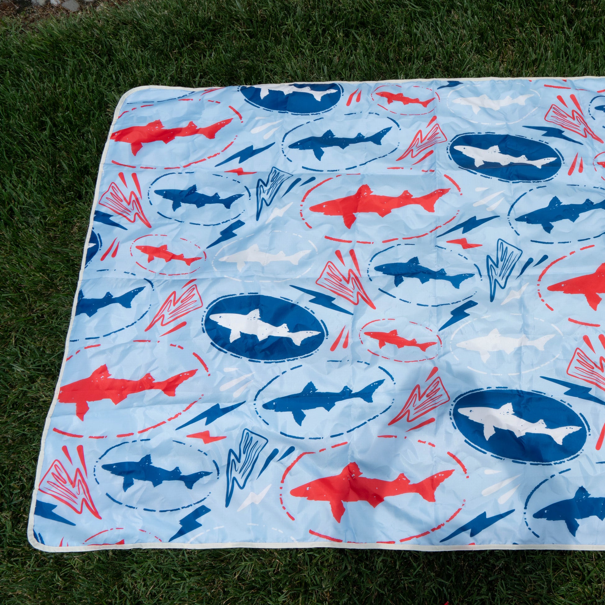Spread out on green grass, this picnic blanket with blue and red sharks, blue circles, and red lightning bolts is perfect for the Merchtank Drink Summer Louder Festival Kit or as a must-have summer festival essential.