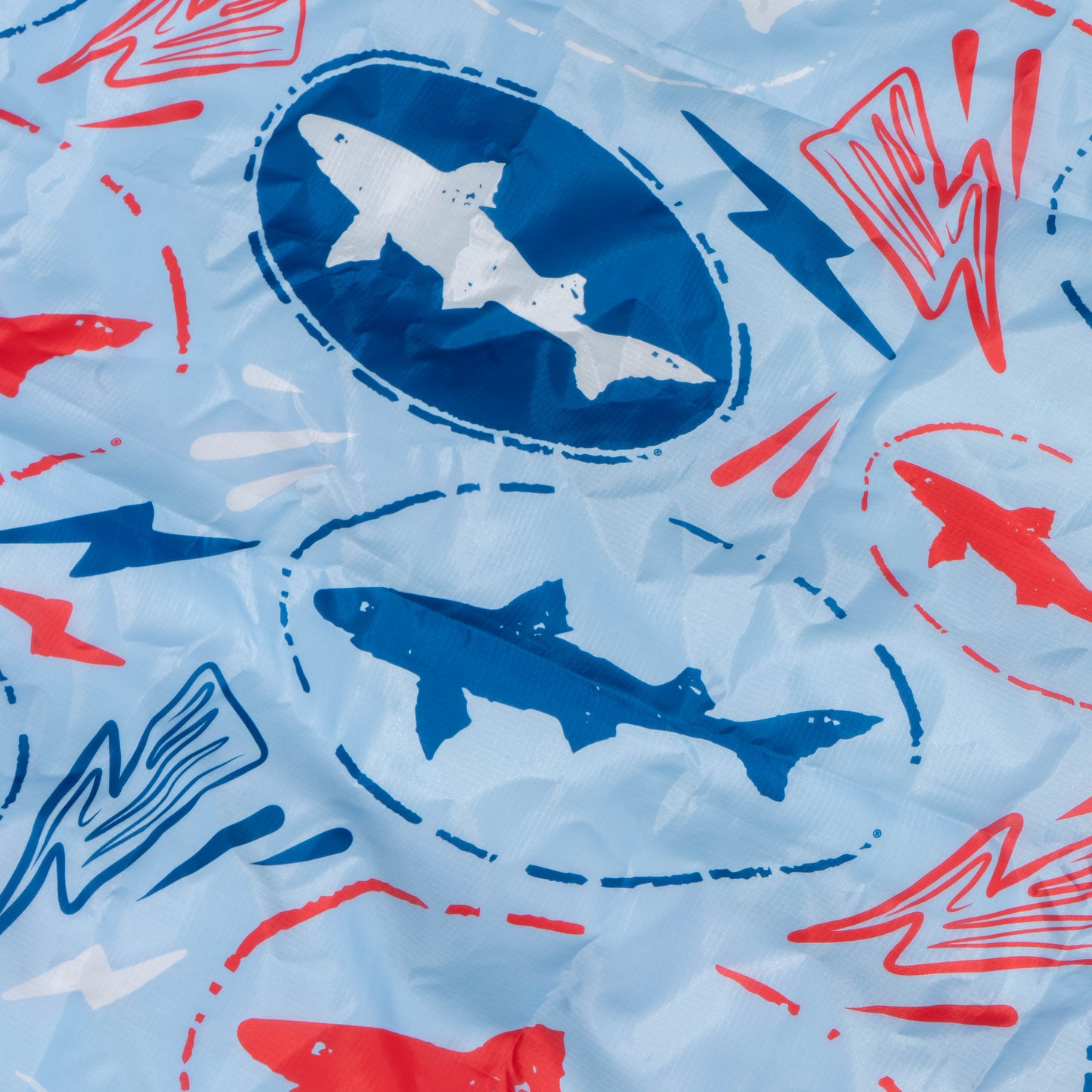 A close-up of the Drink Summer Louder Festival Kit fabric by Merchtank, featuring blue, white, and red sharks in ovals with red and blue lightning bolts—ideal for festival essentials; fabric shows slight wrinkles.