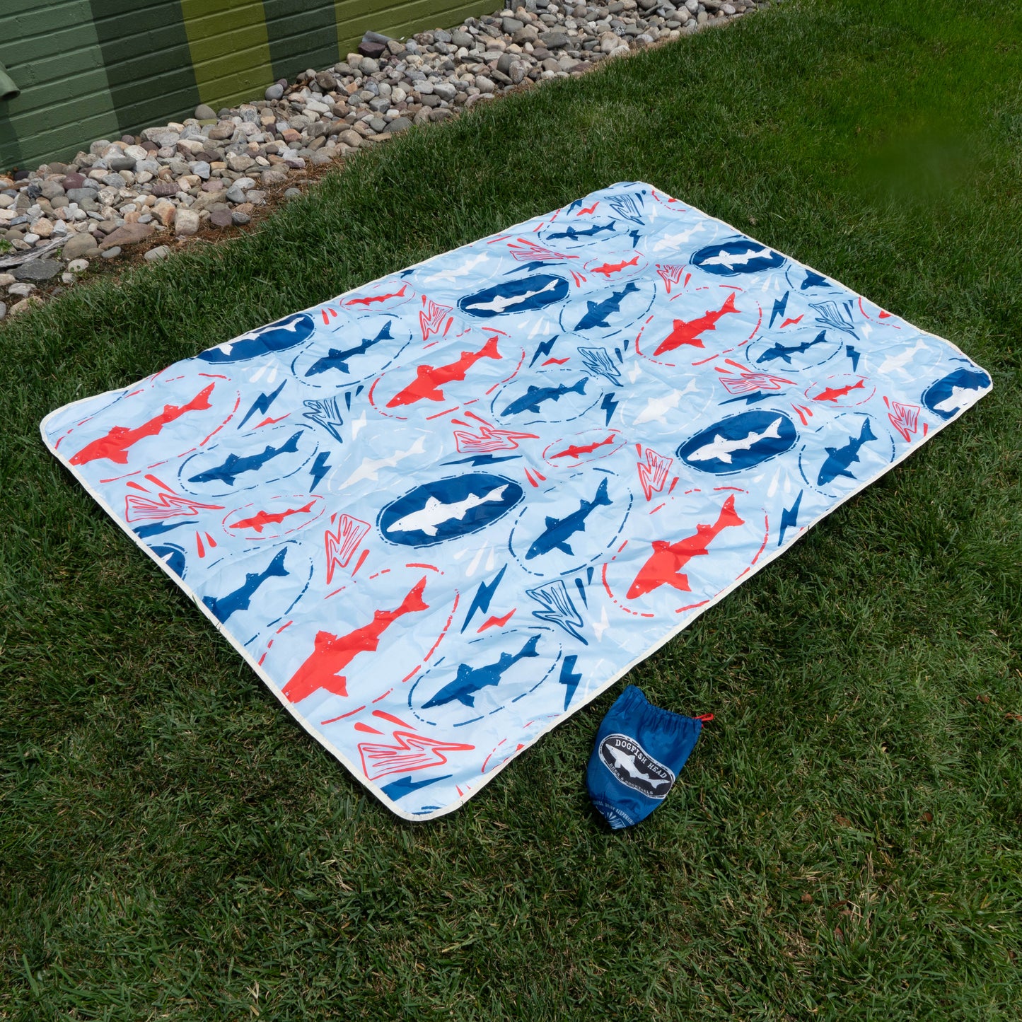 A rectangular picnic blanket with a blue, white, and red shark pattern—part of the Merchtank Drink Summer Louder Festival Kit—lies on green grass. A matching drawstring pouch sits nearby, ideal for carrying your summer festival essentials.