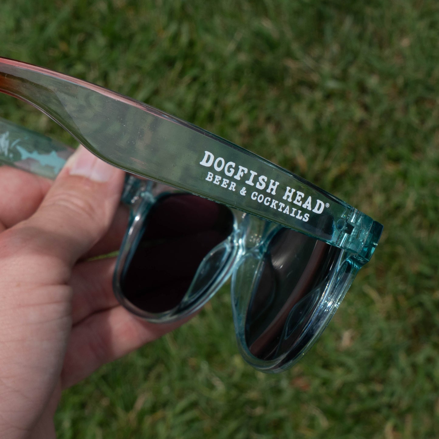 A hand holds teal sunglasses with white "DOGFISH HEAD® BEER & COCKTAILS" lettering against a grassy blur—ultimate summer vibes from the Merchtank Drink Summer Louder Festival Kit.