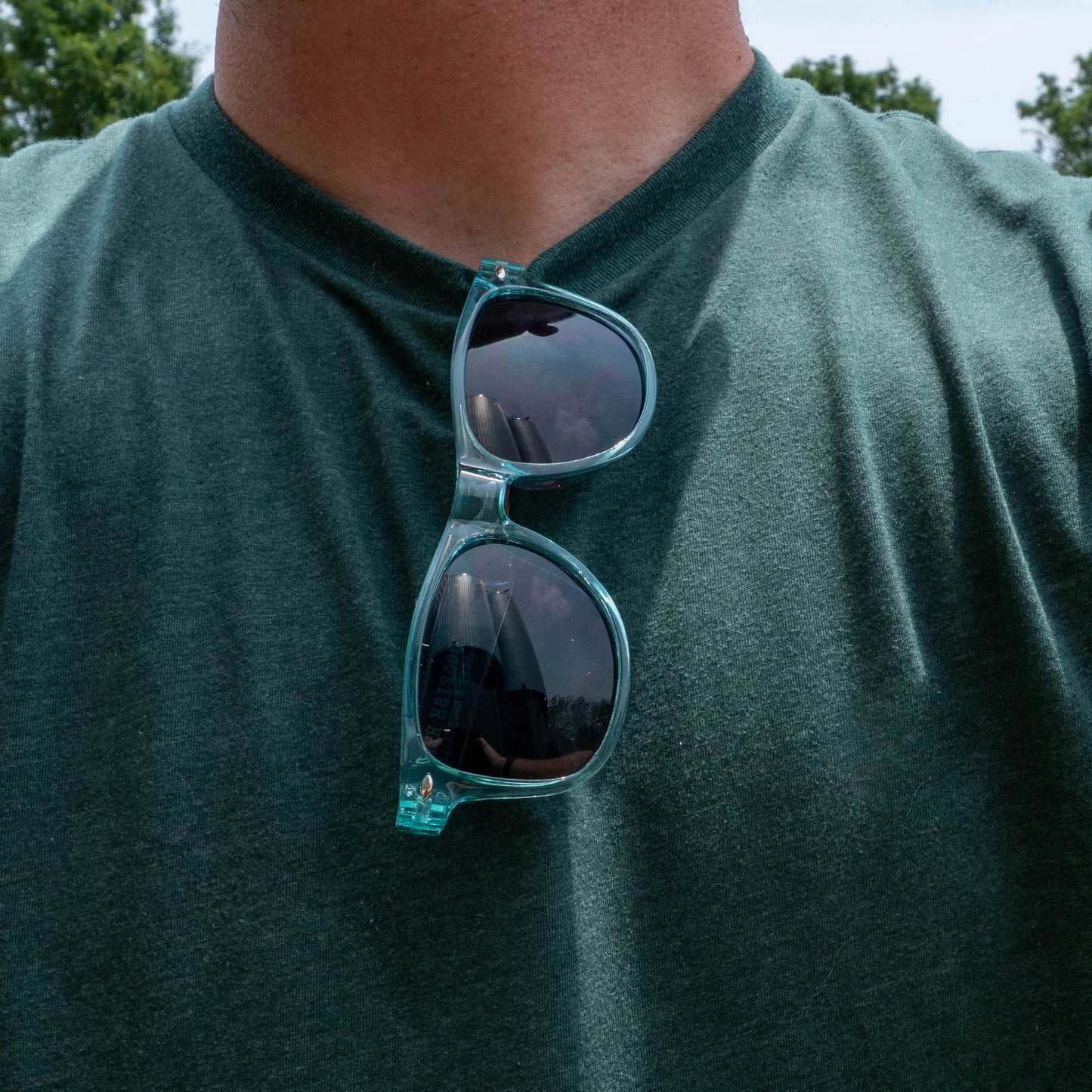 Wearing a green t-shirt and blue sunglasses, someone enjoys the sunny outdoors with trees all around—embracing their Drink Summer Louder attitude and essentials from the Merchtank Drink Summer Louder Festival Kit.