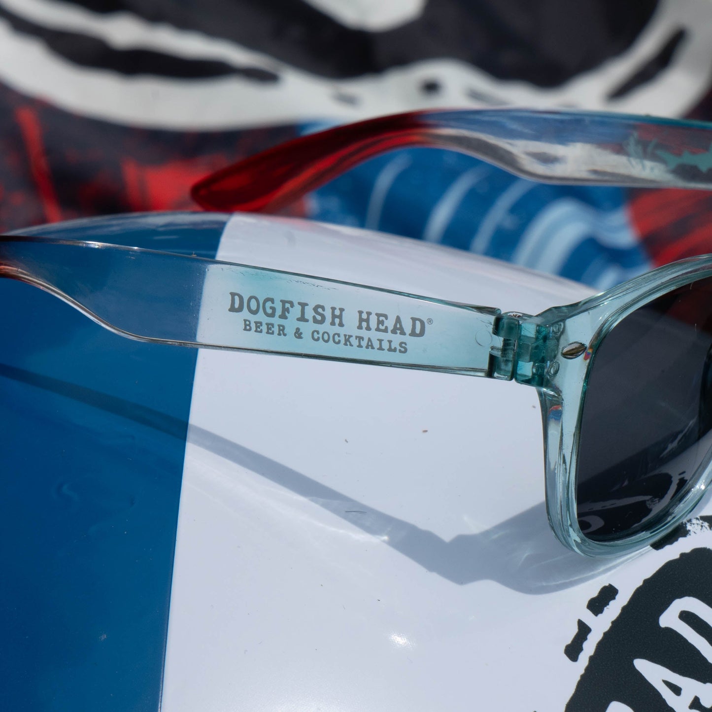 Close-up of blue-tinted sunglasses with DOGFISH HEAD BEER & COCKTAILS on the arm, resting on a colorful surface—part of the Drink Summer Louder Festival Kit by Merchtank, your summer festival essential.
