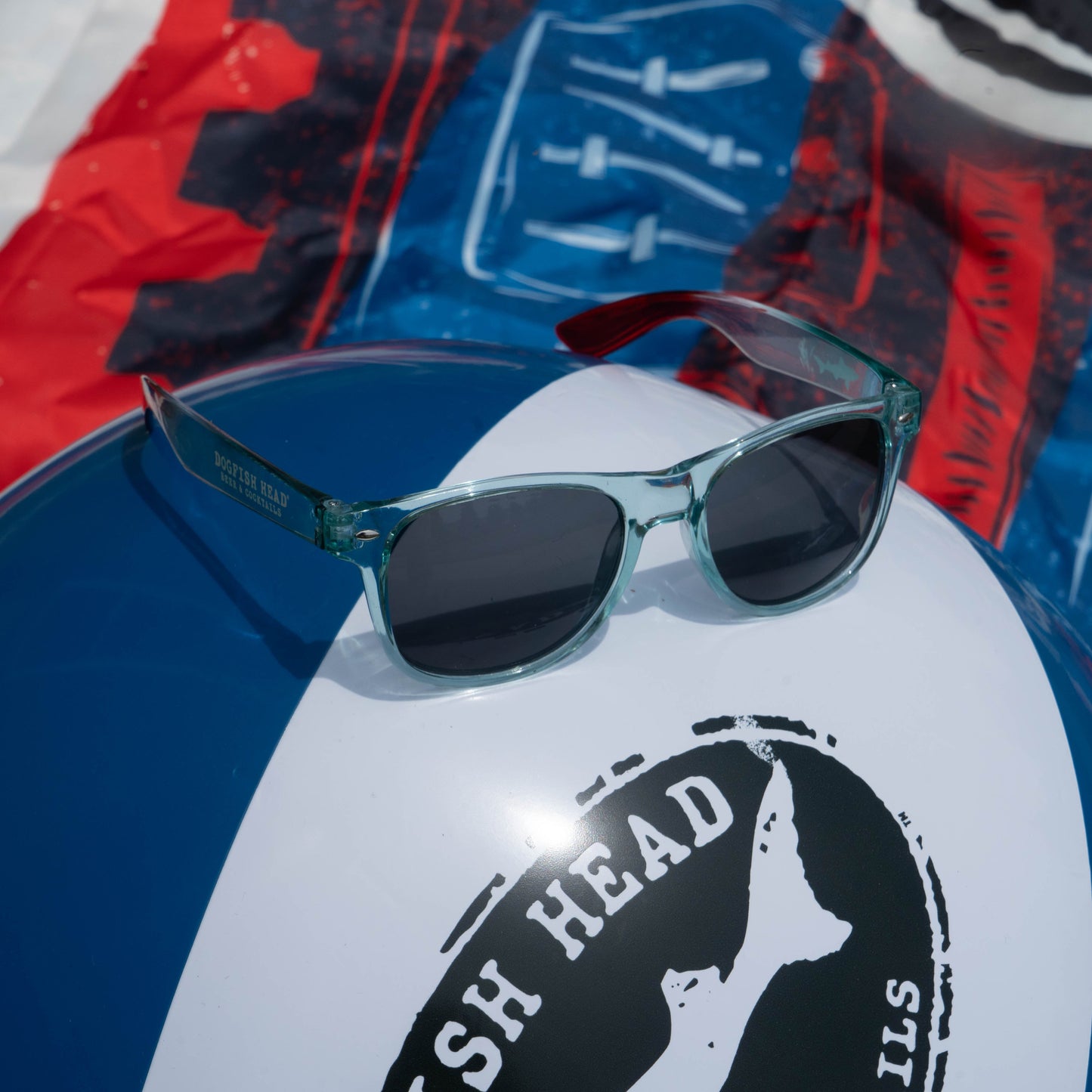 The Drink Summer Louder Festival Kit by Merchtank features translucent turquoise sunglasses resting on a blue and white beach ball with the Fish Head logo, set against a vibrant, blurred background—your festival must-have.