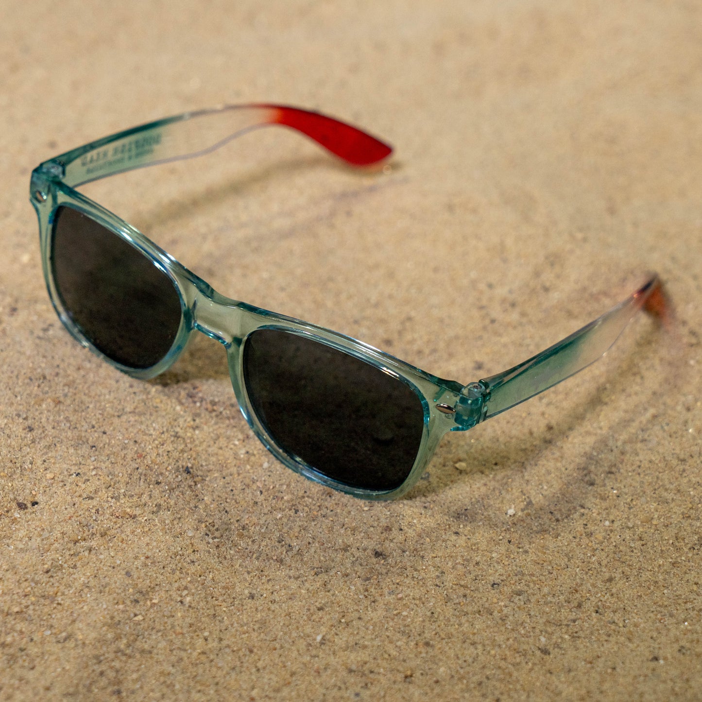The Drink Summer Louder Festival Kit by Merchtank features teal sunglasses with dark lenses and red-tipped arms—essential summer gear for festival goers ready to stand out on sandy grounds.