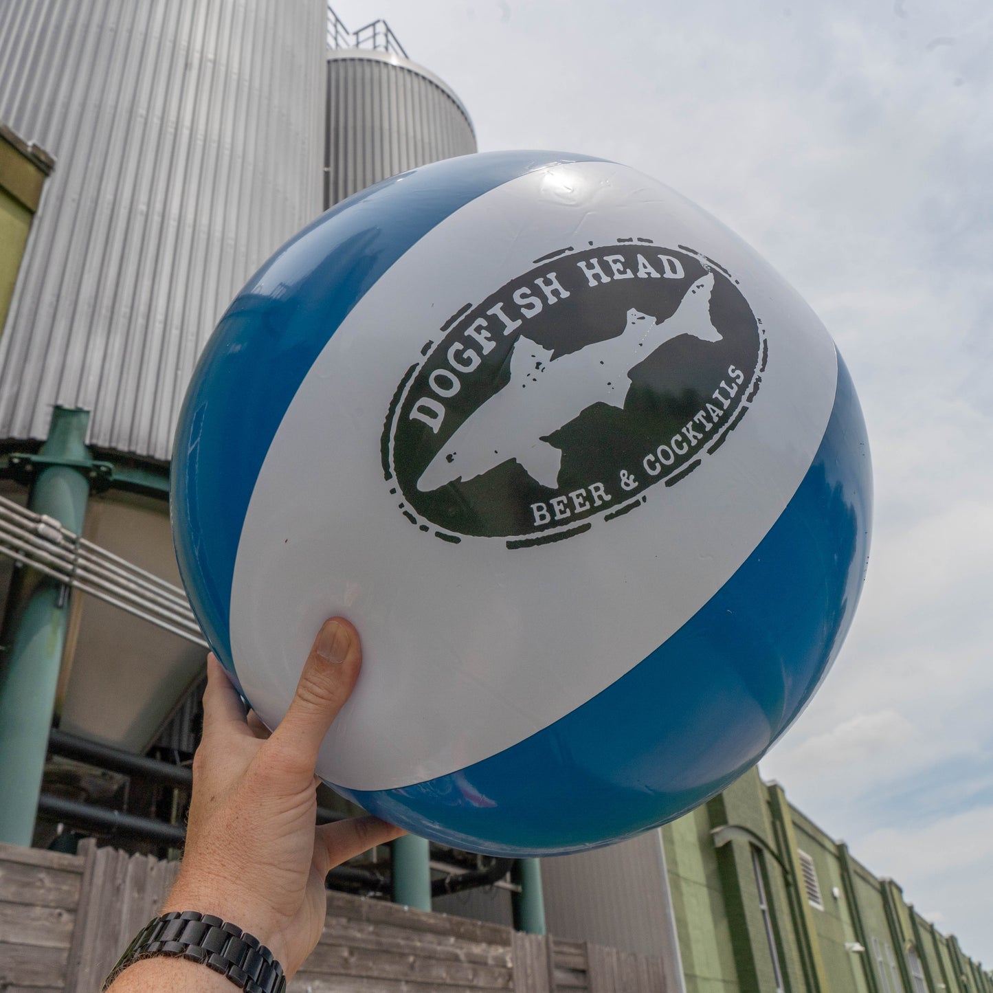 A hand holds up a blue and white beach ball from the Drink Summer Louder Festival Kit by Merchtank in front of brewery tanks under a partly cloudy sky, highlighting a must-have festival essential.