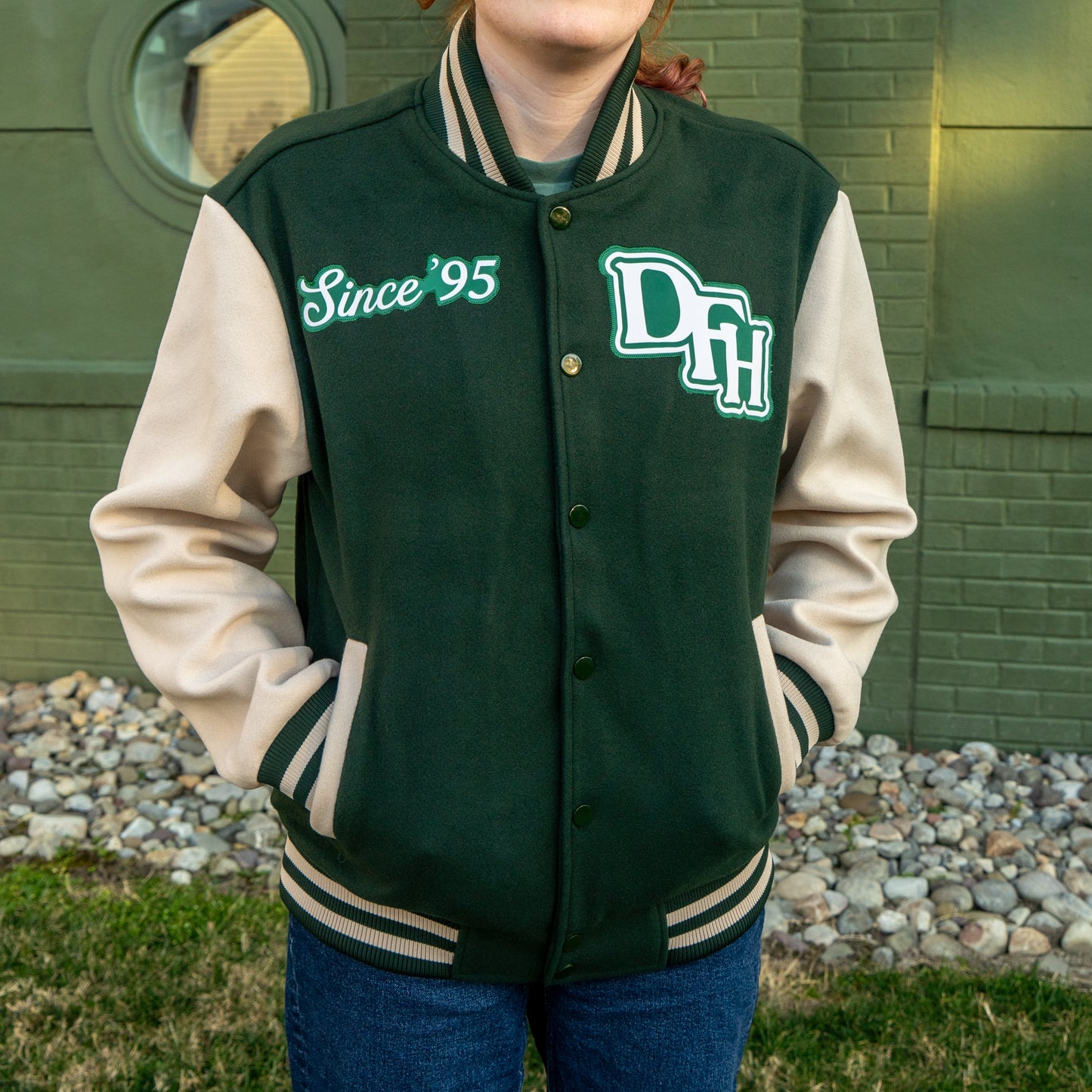 Person wearing a green and beige varsity jacket with 'Since '95' and 'DH' text, standing in front of a brick wall.