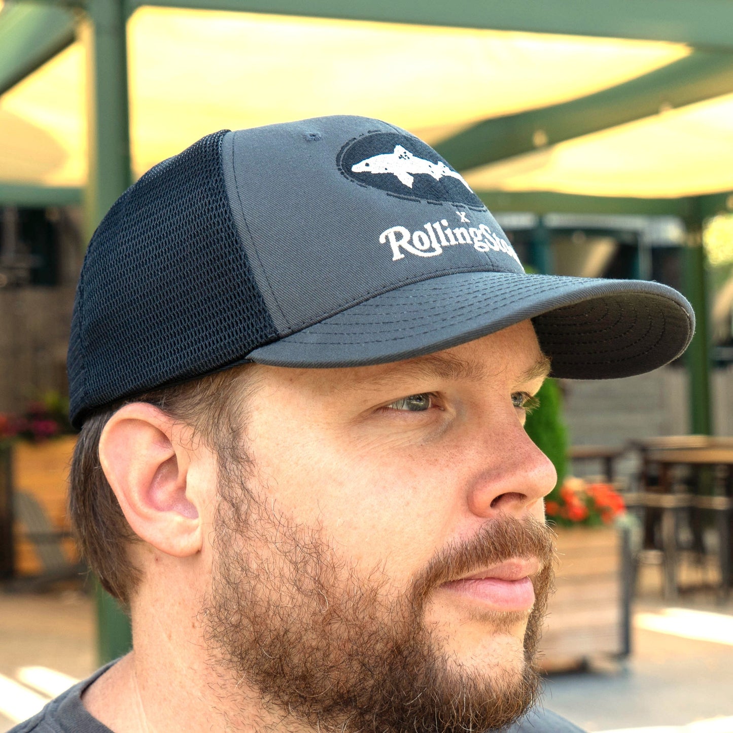 A bearded man outdoors near tables and potted plants wears a Unionwear Dogfish Head x Rolling Stone Trucker Hat, a gray and black snapback with a fish design and embroidered text.