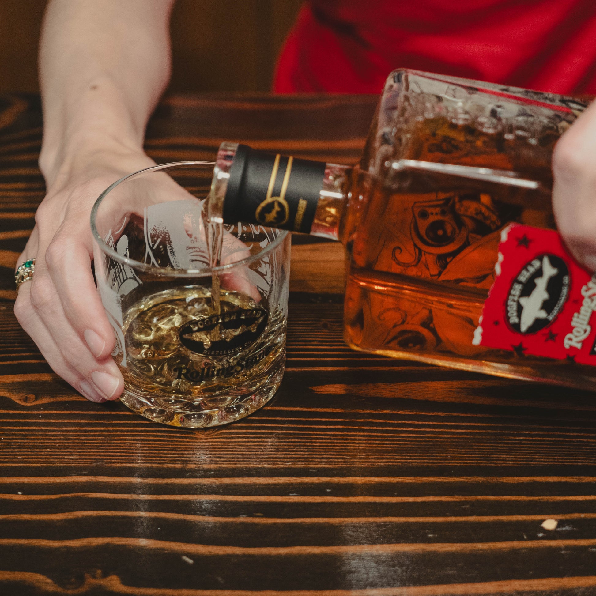 A person pours whiskey from a square bottle into a Dogfish Head x Rolling Stone Whiskey Glass by Grandstand Glassware & Apparel on a wooden table, using one hand to steady the glass, celebrating 250 years of American music.