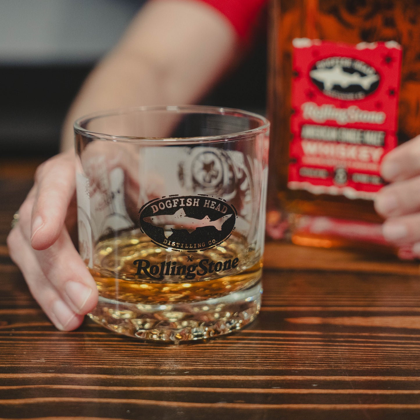A hand holds the Dogfish Head x Rolling Stone Whiskey Glass by Grandstand Glassware & Apparel, celebrating American music; in the background, a matching whiskey bottle with a red label rests on a wooden table.
