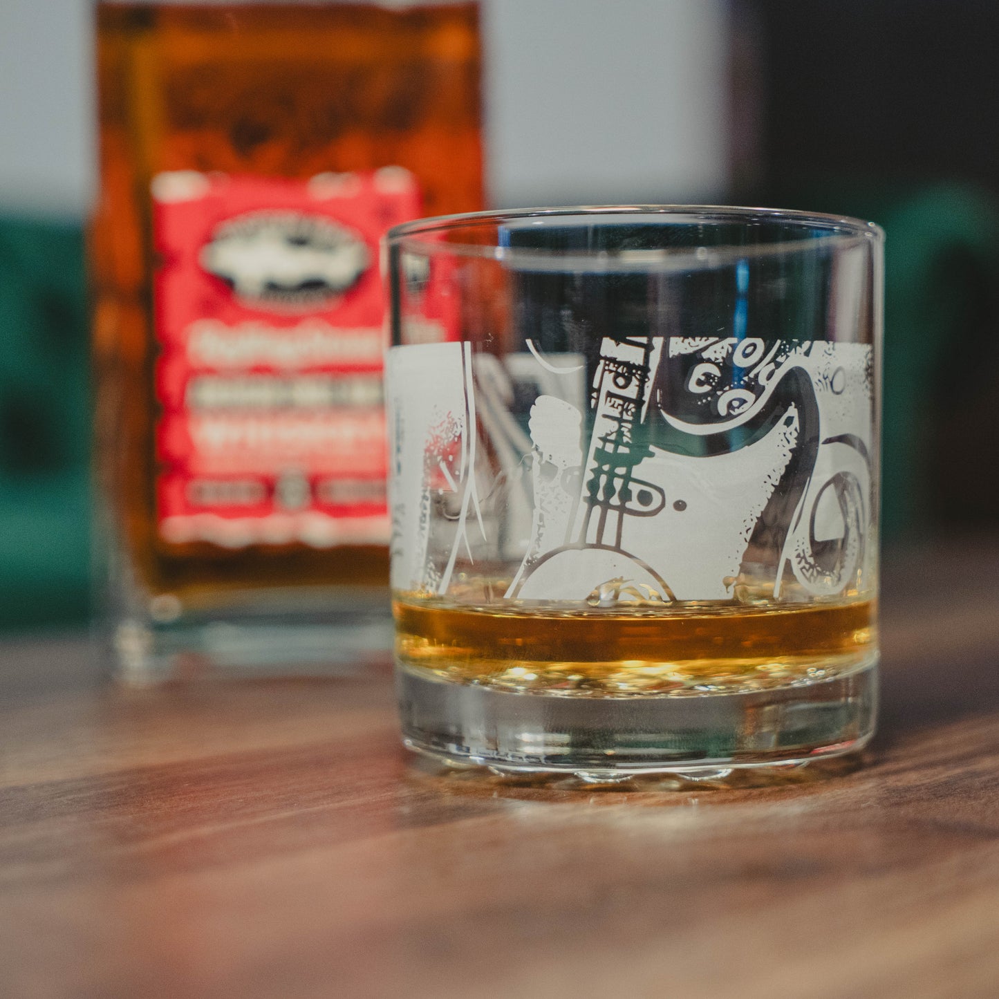 The Dogfish Head x Rolling Stone Whiskey Glass by Grandstand Glassware & Apparel features a unique pattern and holds amber whiskey on wood, with a blurred bottle in back—celebrating American music and craftsmanship.