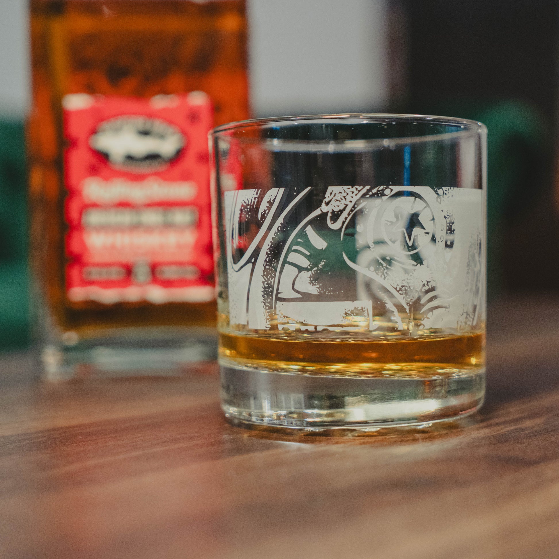 A Dogfish Head x Rolling Stone Whiskey Glass by Grandstand Glassware & Apparel rests on a wooden table, with a blurred bottle in the background—a tribute to American music culture.