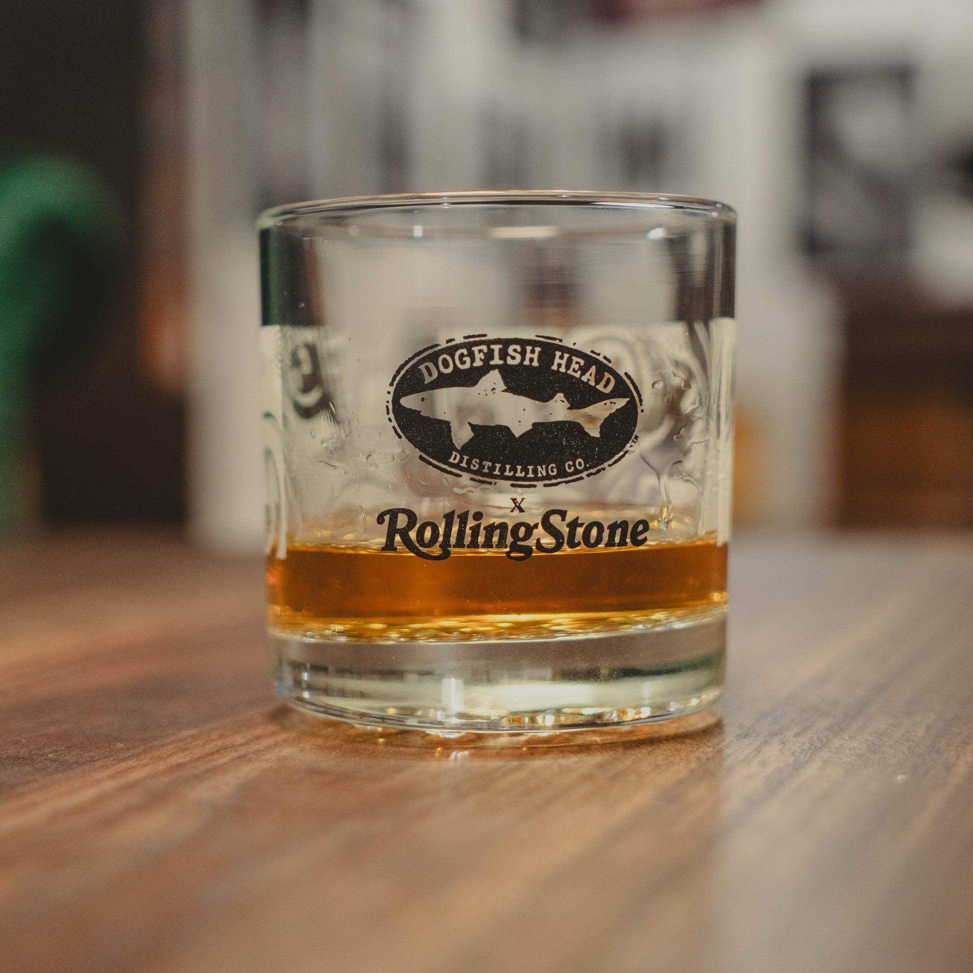 The Dogfish Head x Rolling Stone Whiskey Glass by Grandstand Glassware & Apparel rests on a wooden table, celebrating 250 years of American music in this exclusive collaboration.