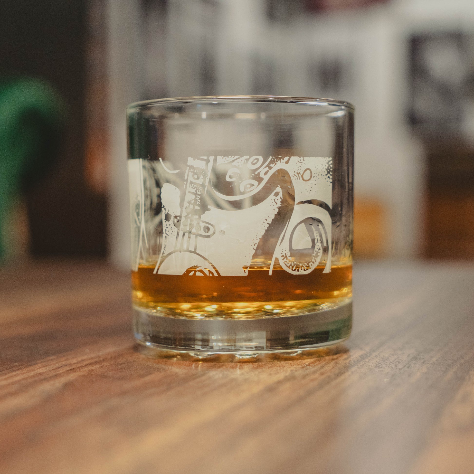 The Dogfish Head x Rolling Stone Whiskey Glass by Grandstand Glassware & Apparel rests on a wooden table, etched with a guitar and musical notes, celebrating 250 years of American music against a softly blurred backdrop.