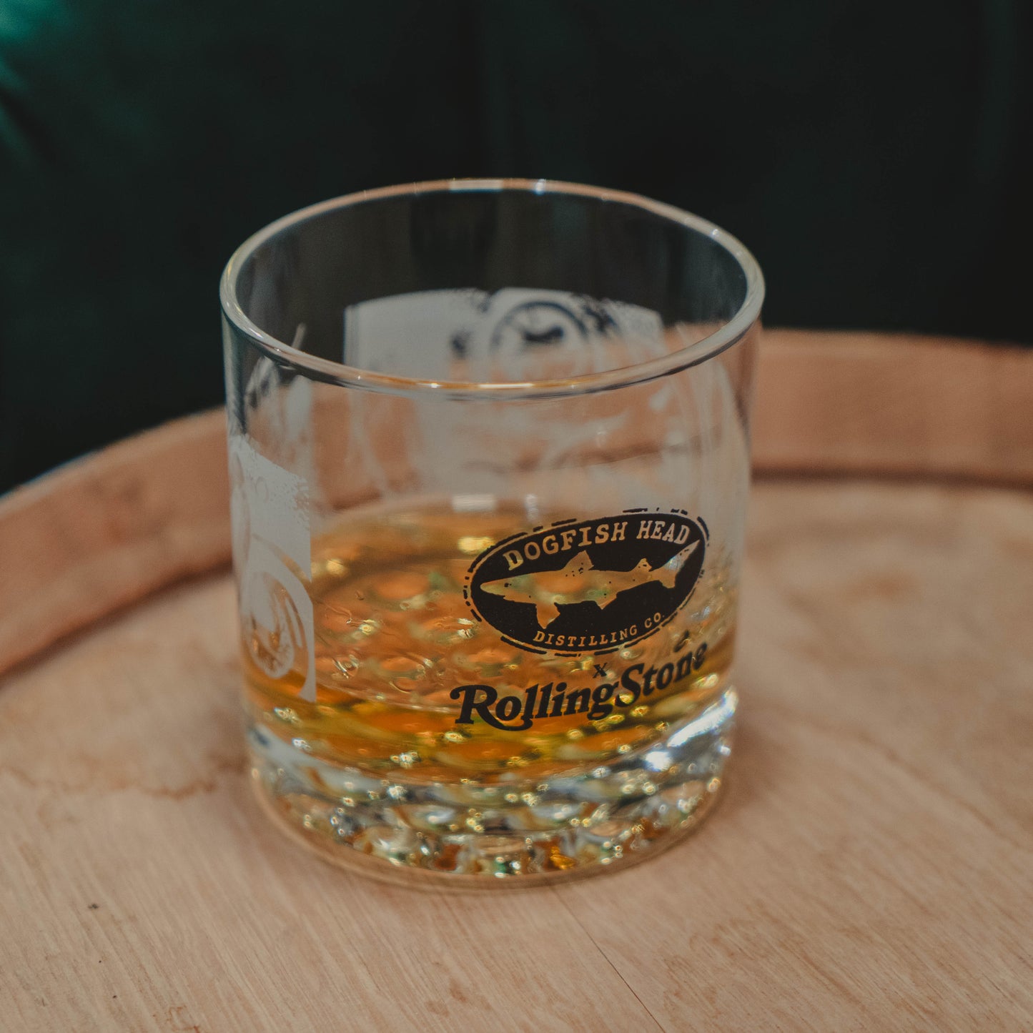 The Dogfish Head x Rolling Stone Whiskey Glass by Grandstand Glassware & Apparel rests on a wooden surface, honoring American music and fine spirits with exclusive branded style.