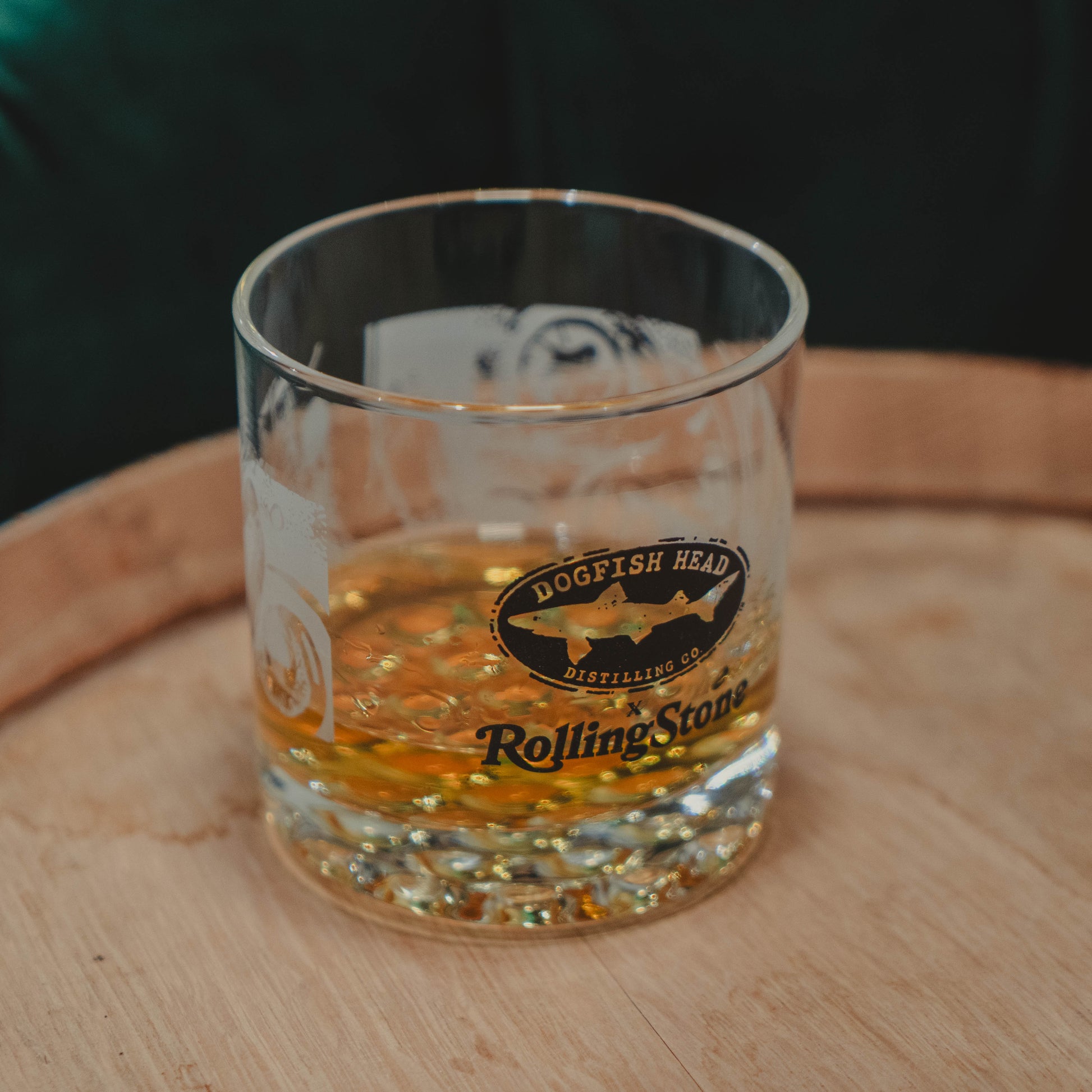 The Dogfish Head x Rolling Stone Whiskey Glass by Grandstand Glassware & Apparel rests on a wooden surface, honoring American music and fine spirits with exclusive branded style.
