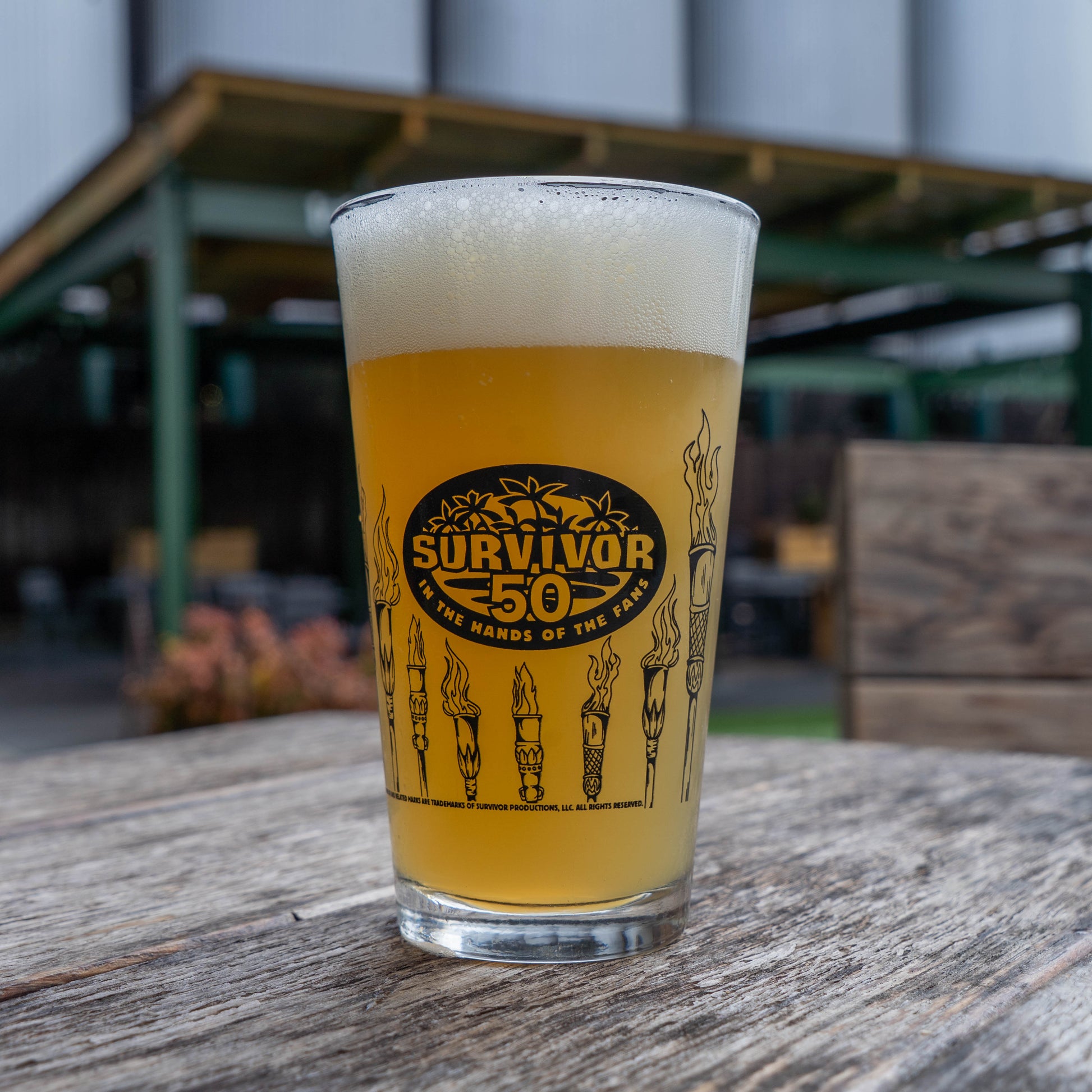A pint glass from the Dogfish Head x Survivor 50 Bundle by Grandstand Glassware & Apparel sits on a wooden table outdoors. This exclusive merch-only collectible is perfect for Survivor fans. A blurred patio area appears in the background.