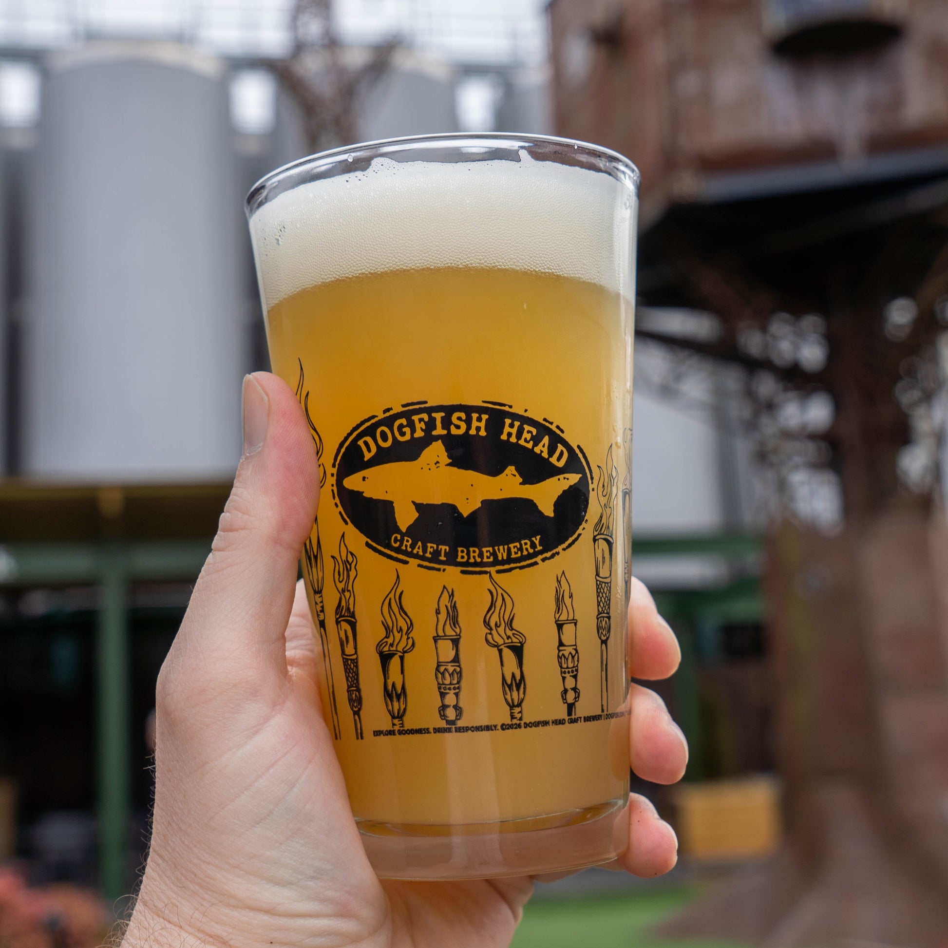 A hand holds a hazy beer in a pint glass with the Dogfish Head logo and torch from the Dogfish Head x Survivor 50 Bundle (MERCH ONLY) by Grandstand Glassware & Apparel; brewery tanks blur in the background.
