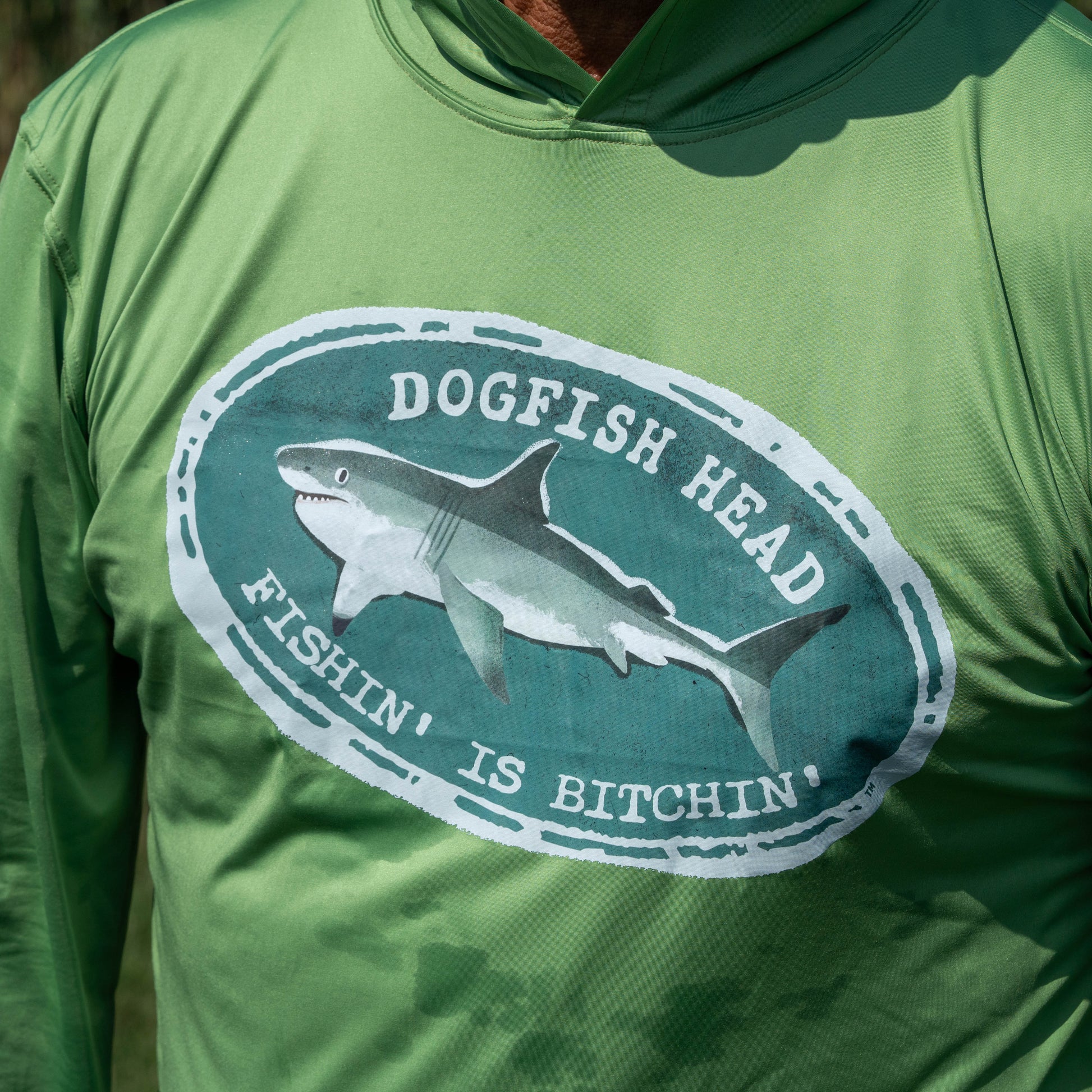 A person wearing the Boathouse Apparel Dogfish Head x Boathouse Fishin' is Bitchin' Sun Pullover Hoodie, a green moisture-wicking shirt featuring an oval logo with a shark and the words "Dogfish Head Fishin' Is Bitchin'" from the collaborative collection.