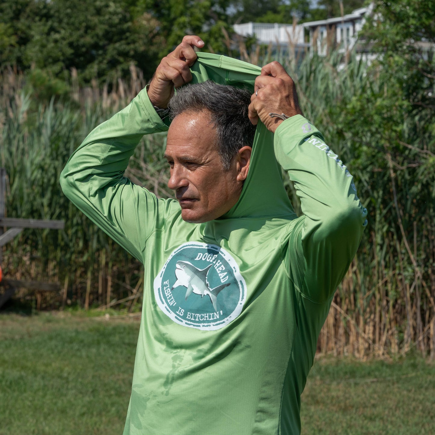 A man in a green Dogfish Head x Boathouse Fishin' is Bitchin' Sun Pullover Hoodie from Boathouse Apparel pulls the hood over his head while standing on grass with tall plants and houses in the background, highlighting this moisture-wicking outdoor performance apparel.