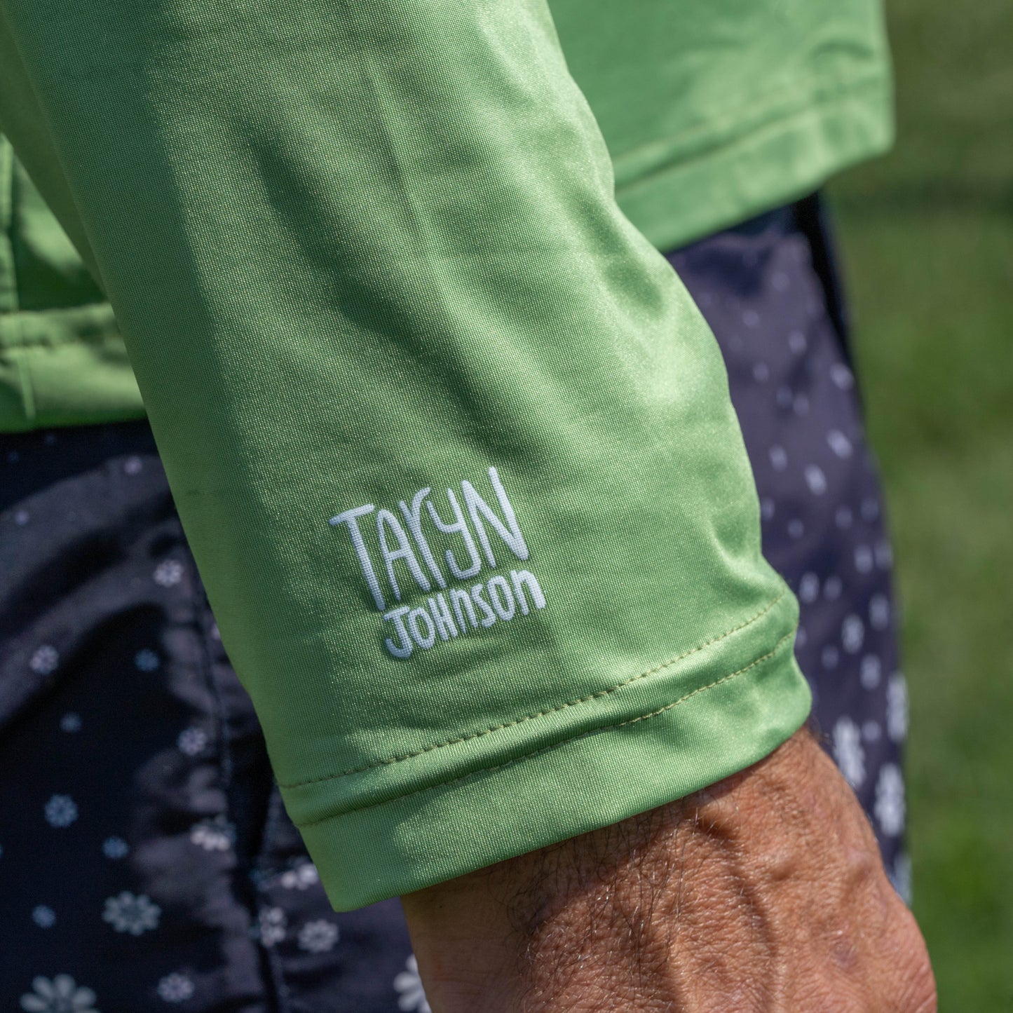 A person wearing a light green Dogfish Head x Boathouse Fishin' is Bitchin' Sun Pullover Hoodie by Boathouse Apparel, featuring long sleeves and TAYJN JOHNSON embroidered on the sleeve, paired with dark shorts with a white floral pattern—showcasing stylish, moisture-wicking outdoor apparel ideal for any Dogfish Head x Boathouse adventure.