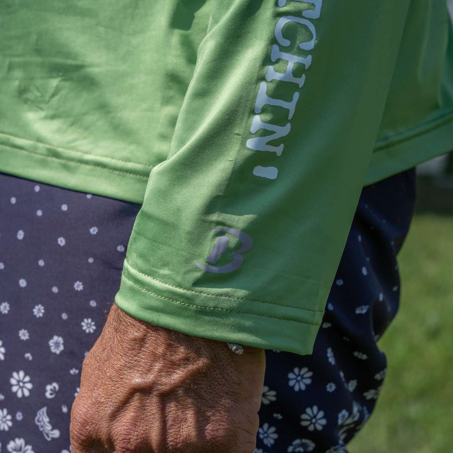 A close-up of a person’s arm wearing a green Boathouse Apparel Dogfish Head x Boathouse Fishin' is Bitchin' Sun Pullover Hoodie with white text, paired with patterned blue shorts. The detailed texture of the person’s hand is visible, and blurred grass appears in the background.