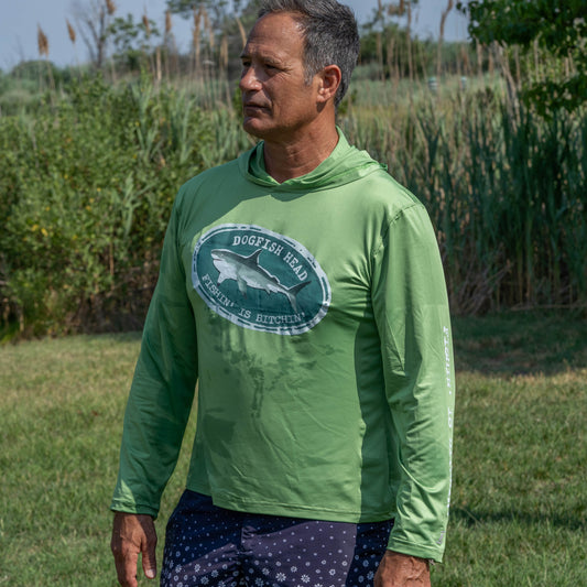 A man wearing a green Boathouse Apparel Dogfish Head x Boathouse Fishin' is Bitchin' Sun Pullover Hoodie and navy shorts with white dots stands outside on grass, with tall plants and trees in the background.