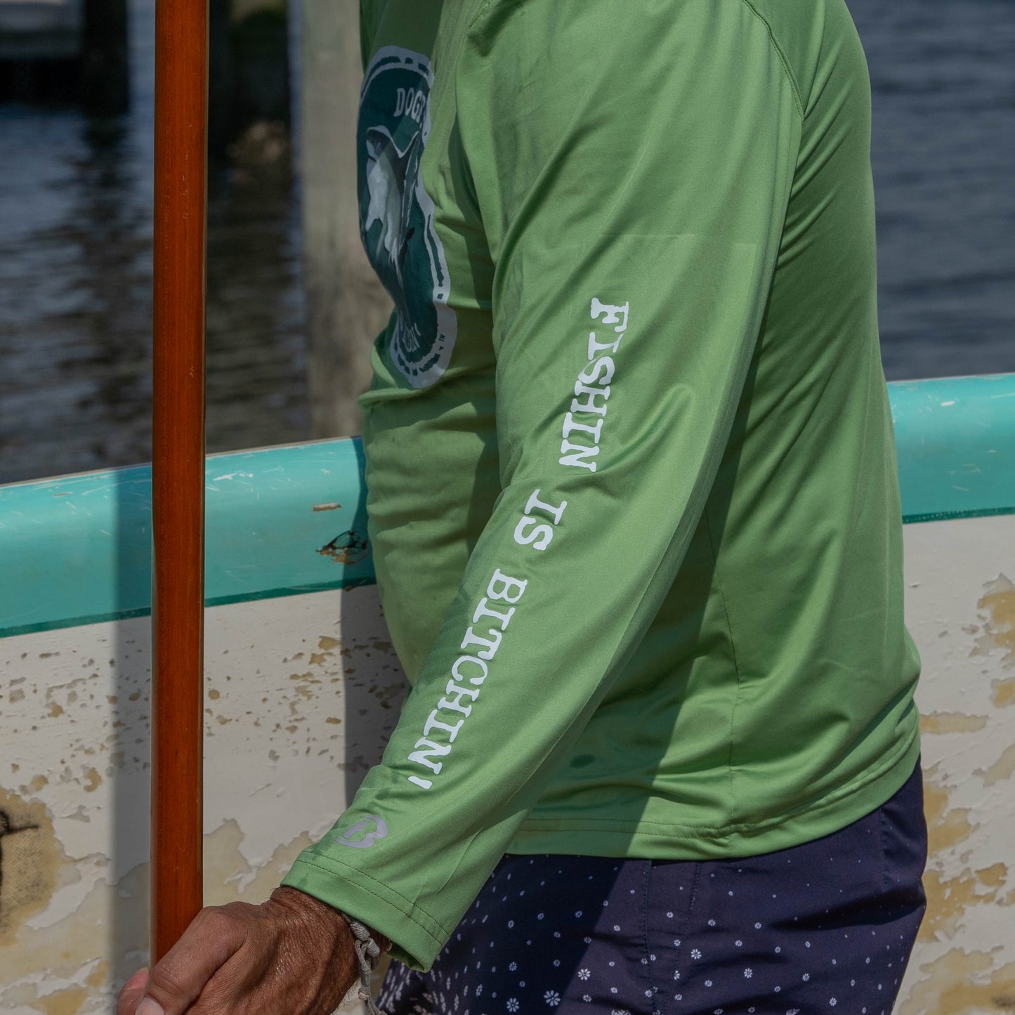 A person wearing the Boathouse Apparel Dogfish Head x Boathouse Fishin' is Bitchin' Sun Pullover Hoodie holds a wooden oar near a weathered boat on the water.