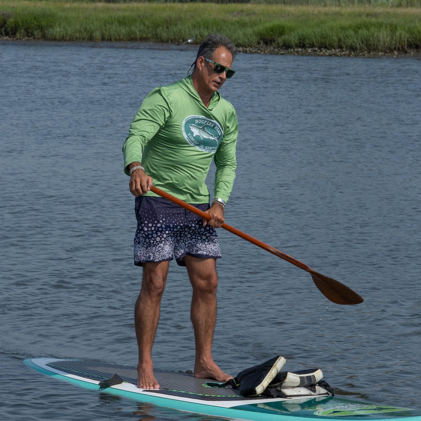 Wearing the Boathouse Apparel Dogfish Head x Boathouse Fishin' is Bitchin' Sun Pullover Hoodie, a person stands on a paddleboard and paddles through calm water, with grass visible in the background.