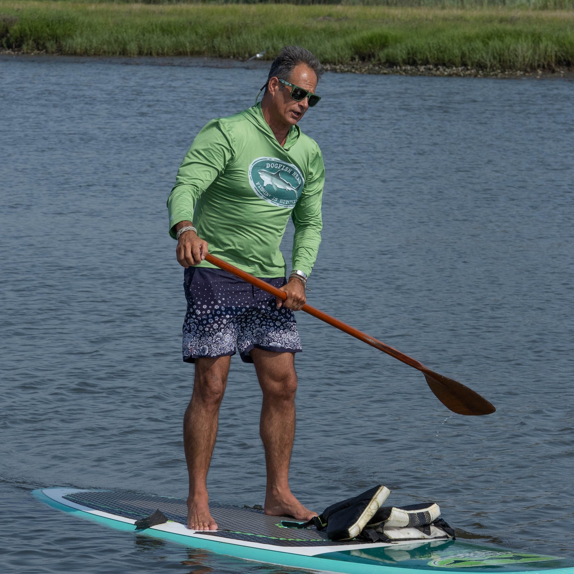 Wearing the Boathouse Apparel Dogfish Head x Boathouse Fishin' is Bitchin' Sun Pullover Hoodie, a person stands on a paddleboard and paddles through calm water, with grass visible in the background.