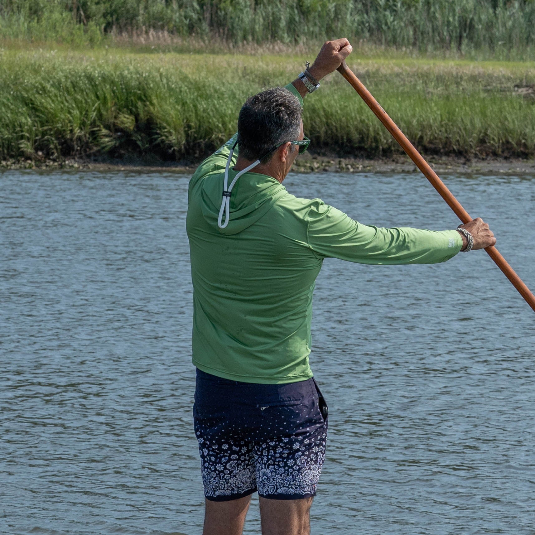 Wearing the Dogfish Head x Boathouse Fishin' is Bitchin' Sun Pullover Hoodie by Boathouse Apparel, a person stands on a paddleboard, holding a paddle over a calm river with tall grass in the background.
