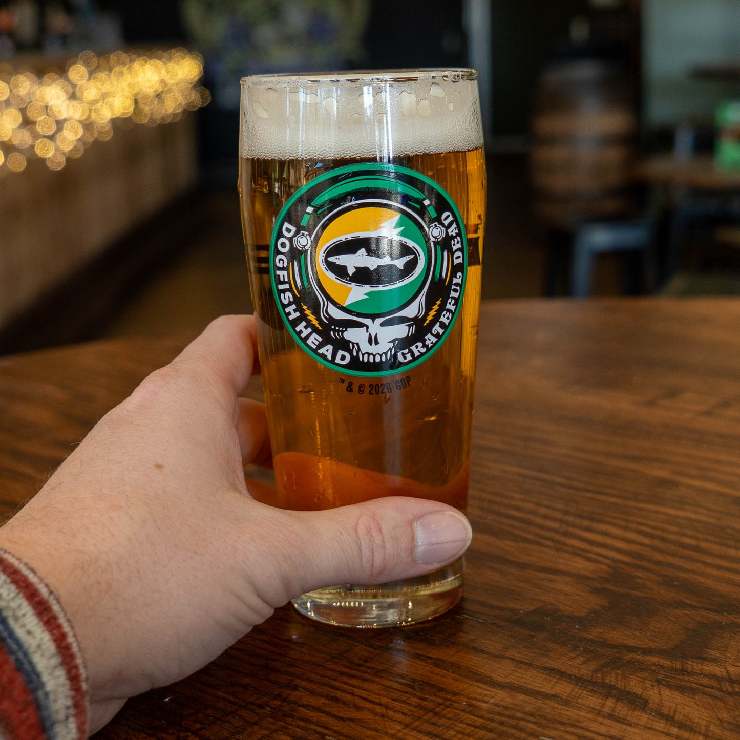 A hand holds a Dogfish Head x Grateful Dead Citrus Daydream Glass by Grandstand Glassware & Apparel on a wooden table; the bar background has string lights and blurred seating.