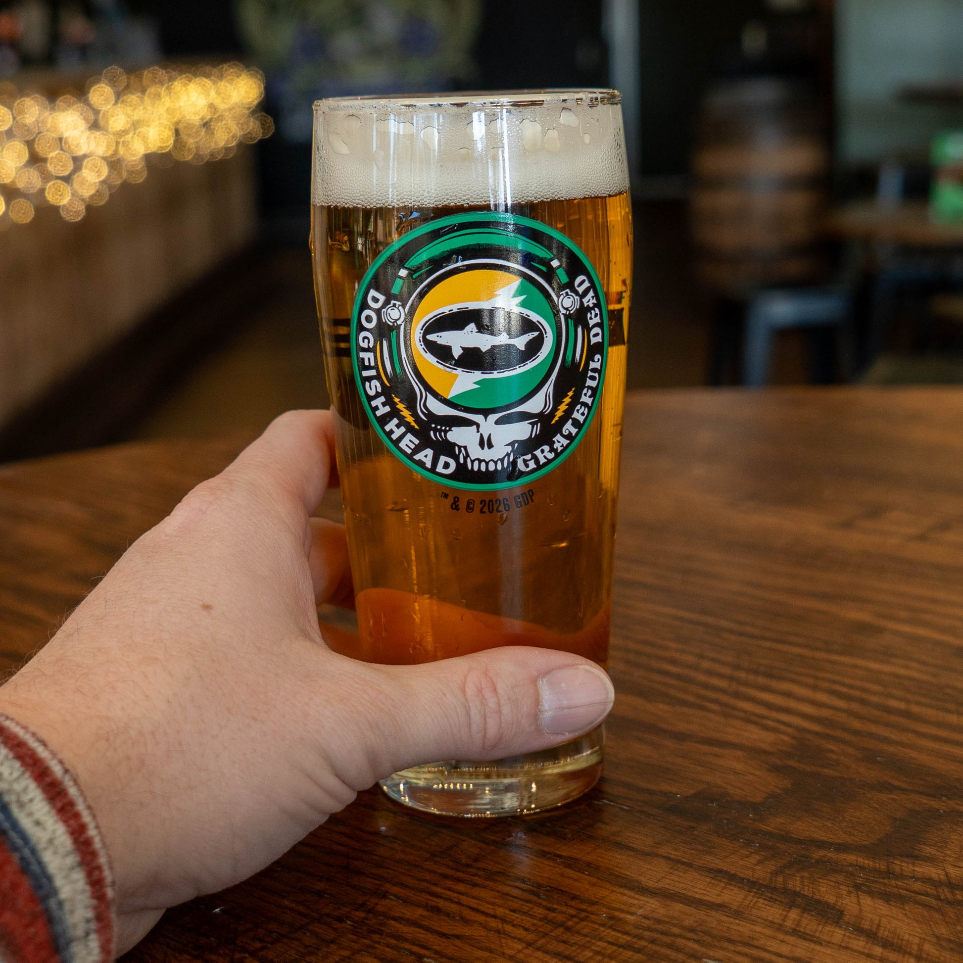A hand holds a Dogfish Head x Grateful Dead Citrus Daydream Glass by Grandstand Glassware & Apparel on a wooden table; the bar background has string lights and blurred seating.