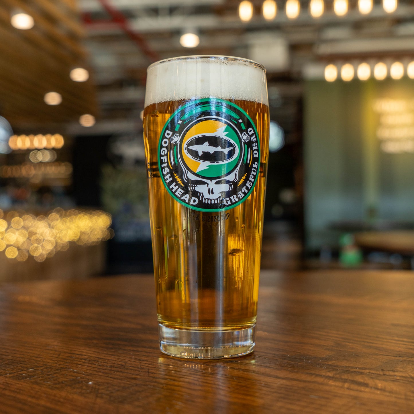 A Dogfish Head x Grateful Dead Citrus Daydream Glass by Grandstand Glassware & Apparel, filled with golden beer, sits on a wooden table. The softly blurred background shows string lights and a bar area.