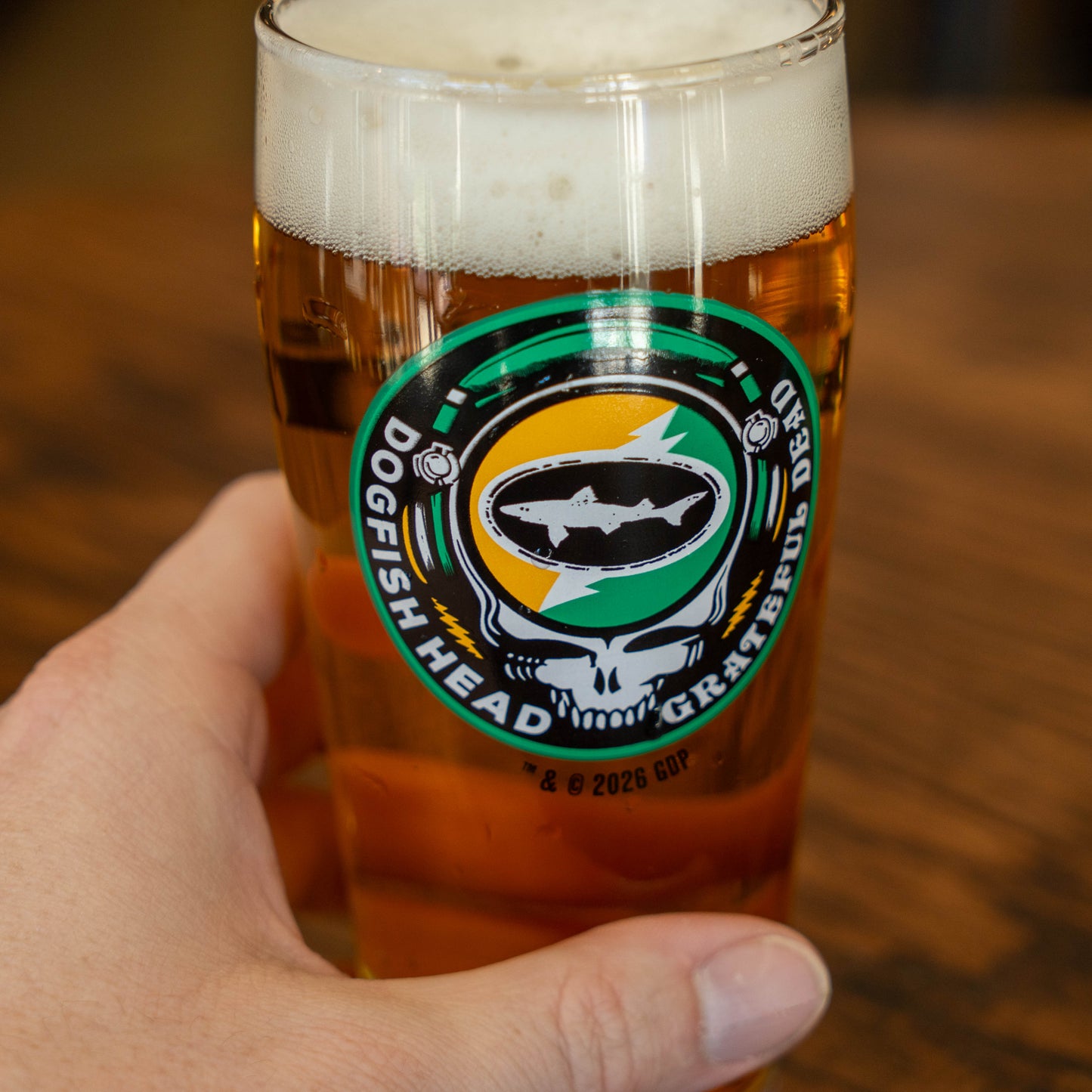 A hand holds the Dogfish Head x Grateful Dead Citrus Daydream Glass from Grandstand Glassware & Apparel, featuring a shark and a skull with a lightning bolt. The pint glass of amber beer rests on a wooden surface.