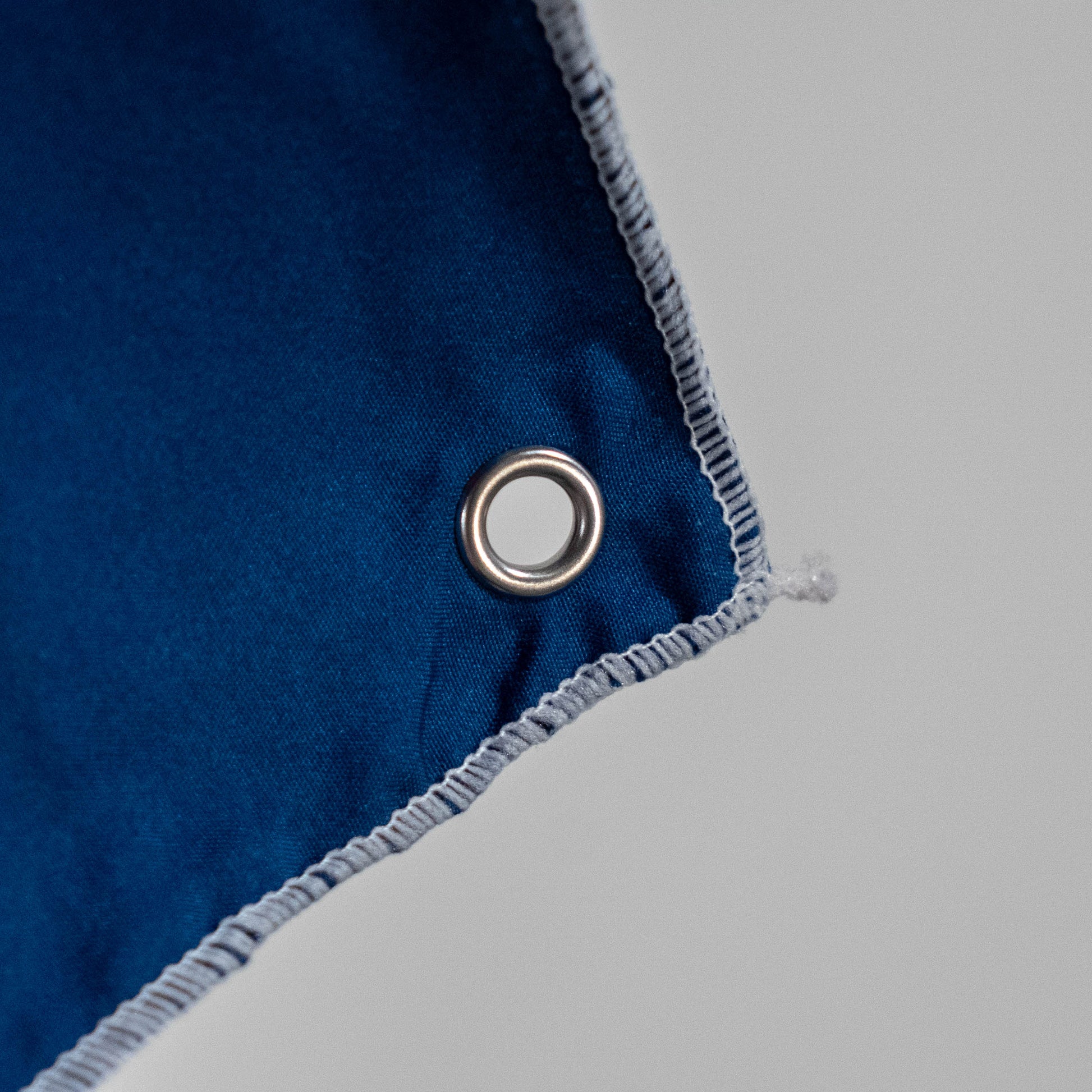 Close-up of the Merchtank Dogfish Head x Grateful Dead Flag Tapestry, showing a blue fabric corner with white stitching and a metal grommet, set against a light grey background.